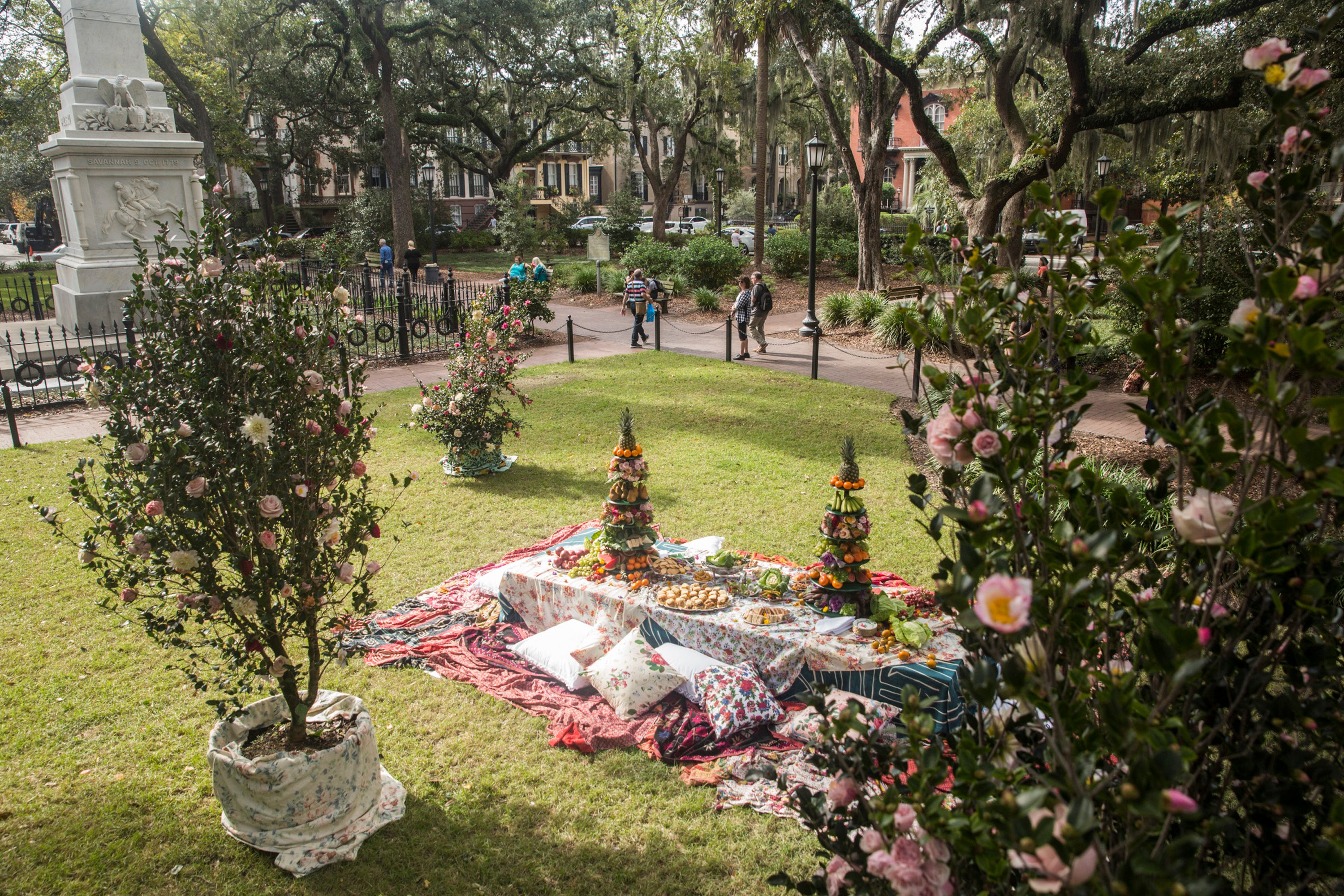 A picnic table set with colorful floral tablecloths and tall platters of produce sits in a Savannah park square surrounded by oak trees, camellia bushes, and historic architecture.