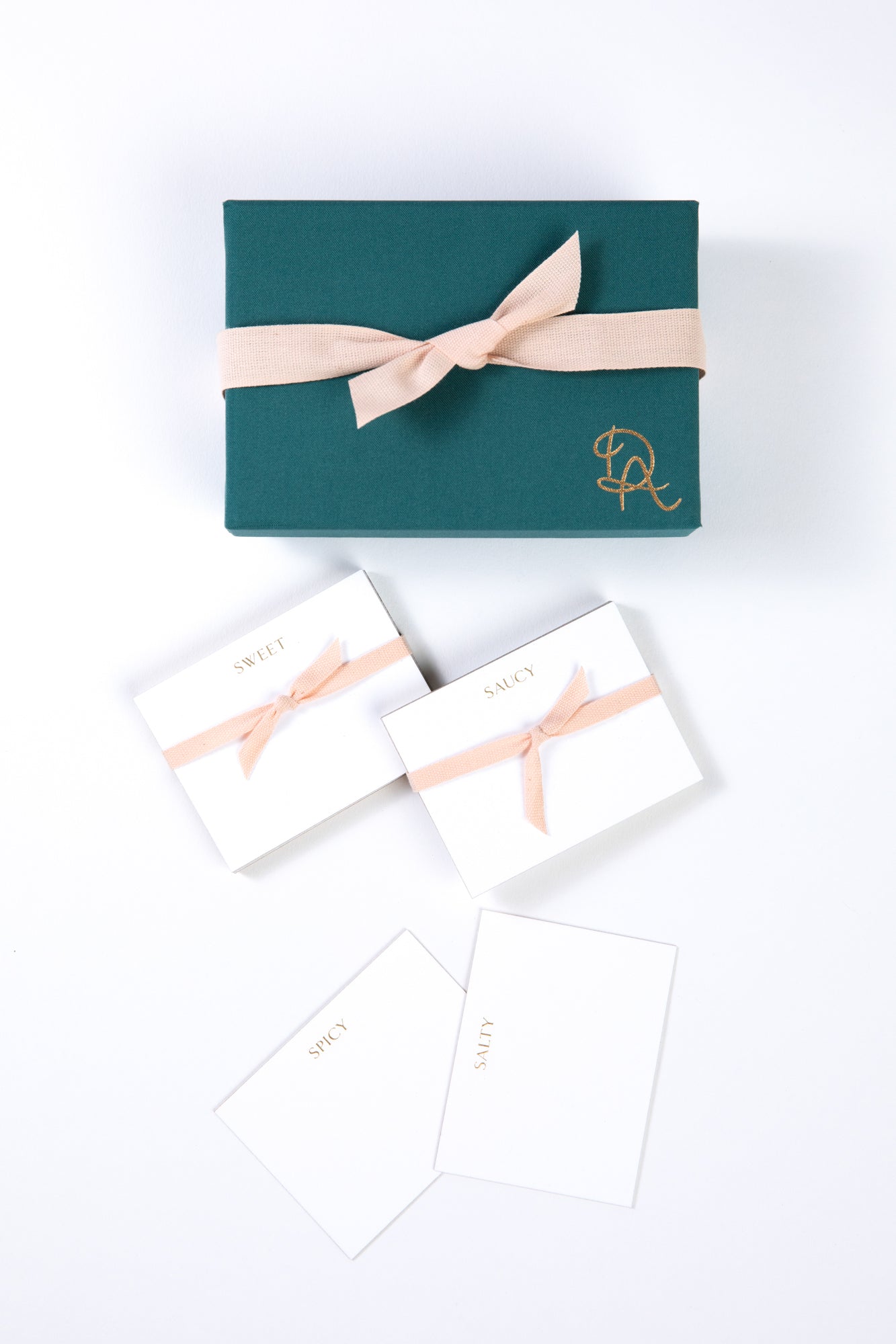 Box and set of white place cards displayed on a white background. Place cards are white with gold engraved lettering across the top featuring a variety of words including "saucy", "salty", "spicy", and "sweet." Four cards with each word are displayed next to a blue box with a gold logo and light pink ribbon.
