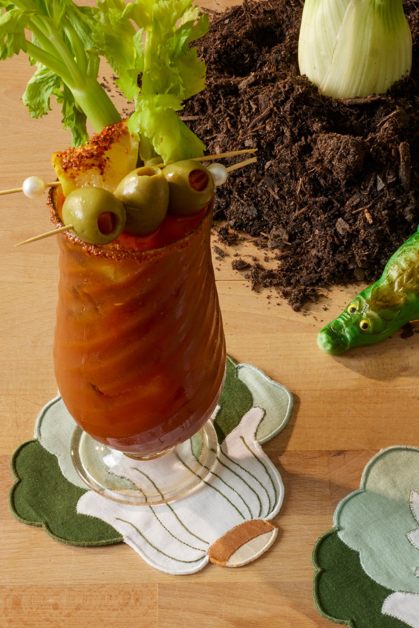 Hand blown glass goblet filled with a bright red bloody mary drink and topped with a seasoned rim, olives, and celery. Bloody mary placed atop a bok choy cocktail napkin. Sitting on wooden tabletop alongside small pile of dirt with toy alligator sneaking into frame.