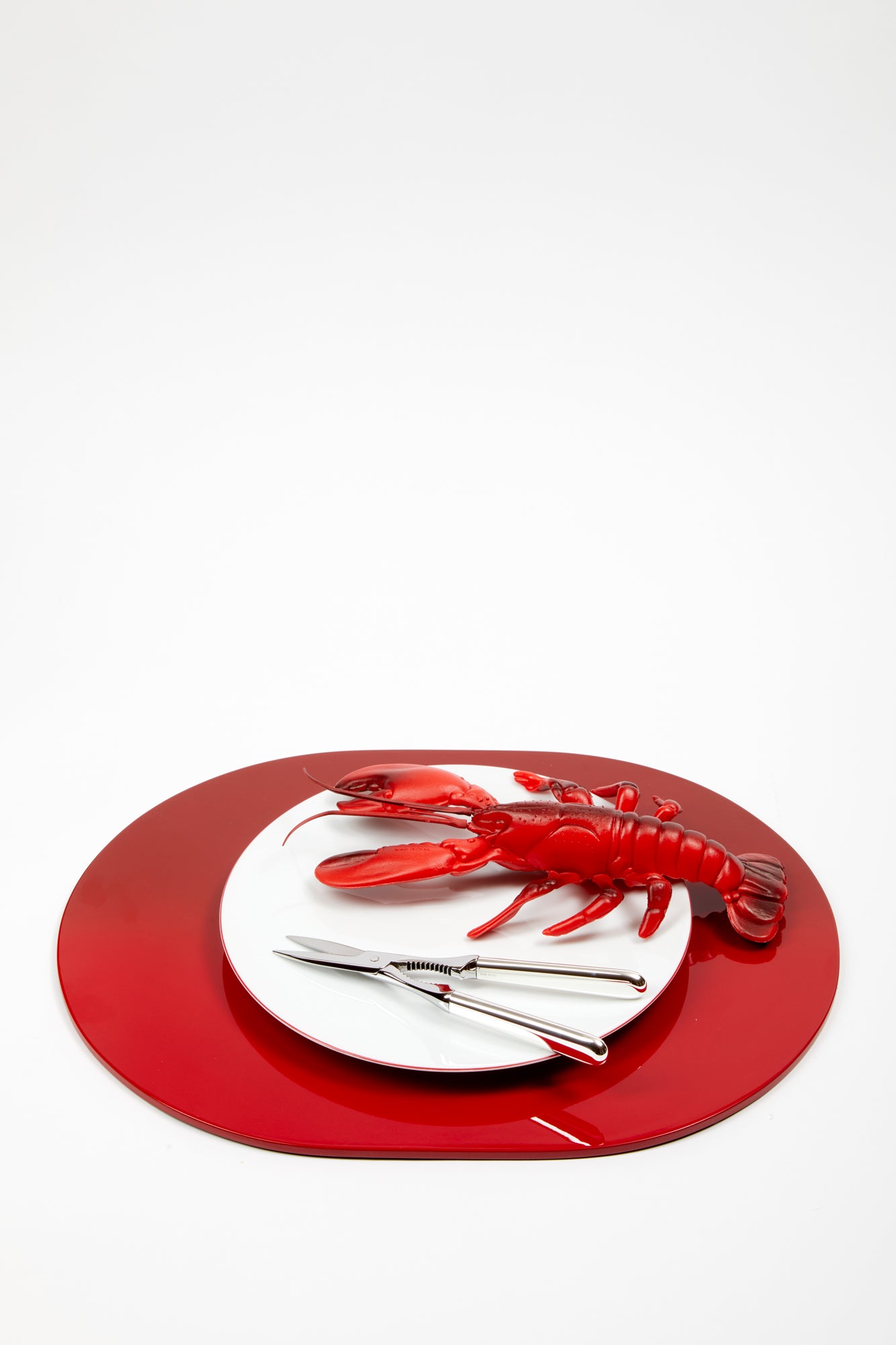 Silver lobster tongs resting aside a fake red lobster, both atop a white plate that is resting on a red lacquer tray.