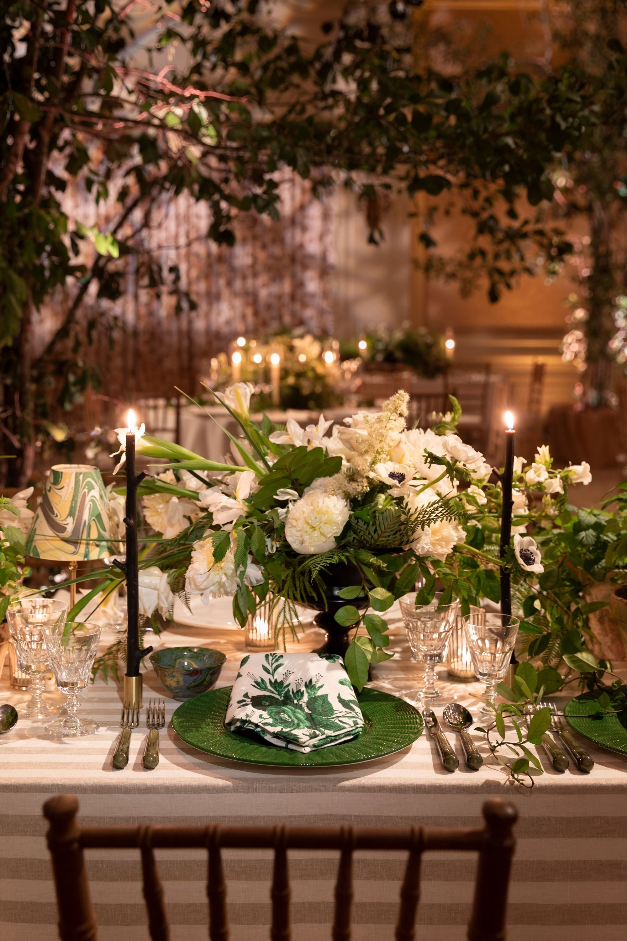 Photograph of a table setting featuring fresh flowers, black stick taper candles in brass candlestick holders, fine silver utensils, "De La Tour" green and white floral napkins, and fine crystal glasses.