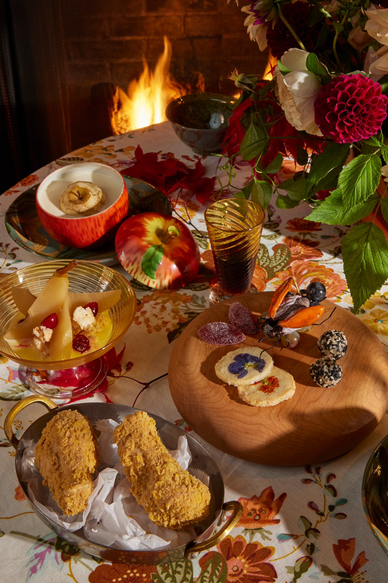 Table setting with variety of decor, including hand blown glass dessert bowl, hand blown glass goblet, wooden passing tray, marbled dish, and more, all atop a floral tablecloth. Complemented by fresh flowers and a crackling fire in the fireplace.