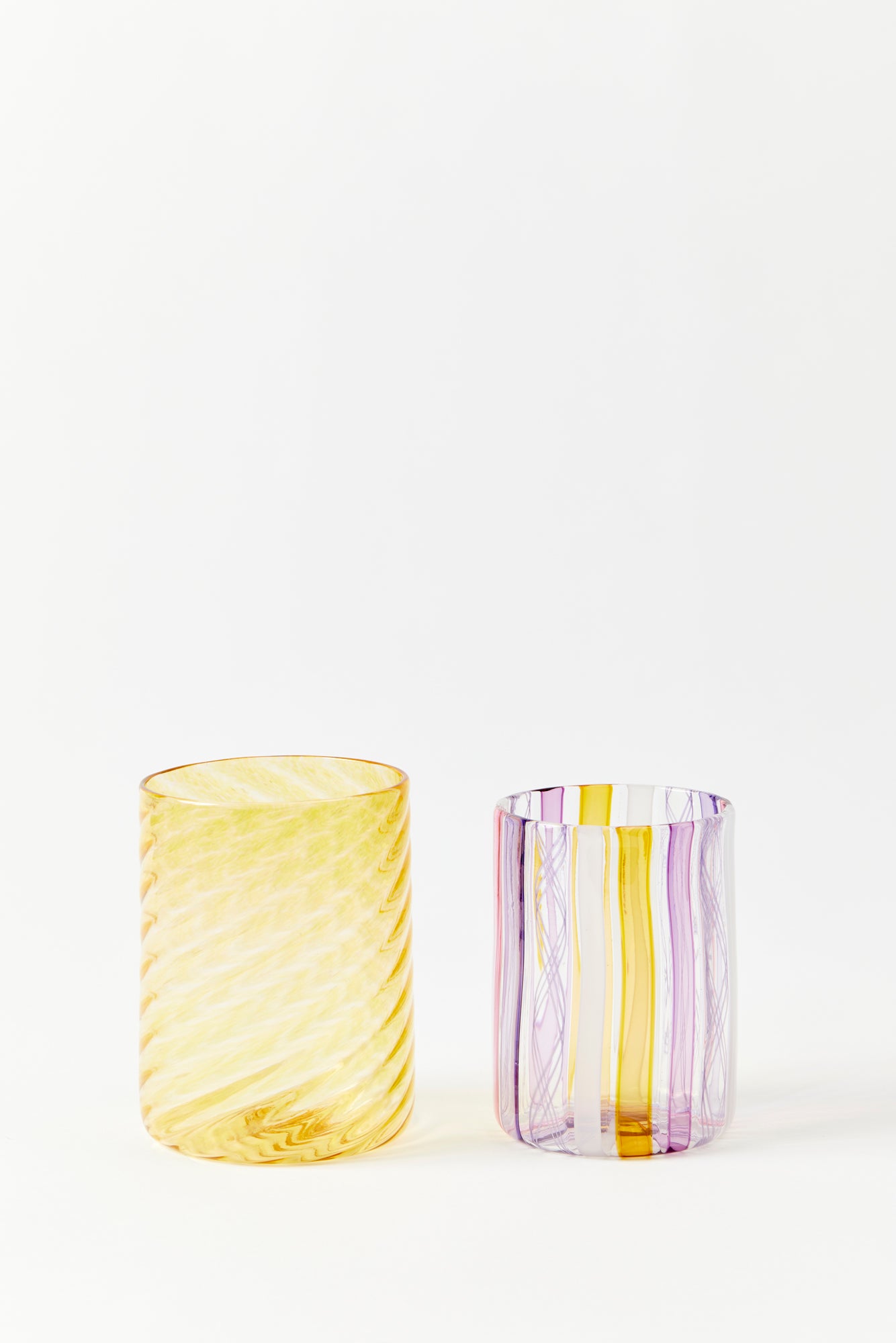 Two hand blown glasses against a white background. On the left is a transparent gold tumbler style glass with a twist pattern. On the right is a tumbler style glass with vertical stripes of pink, yellow, and white.
