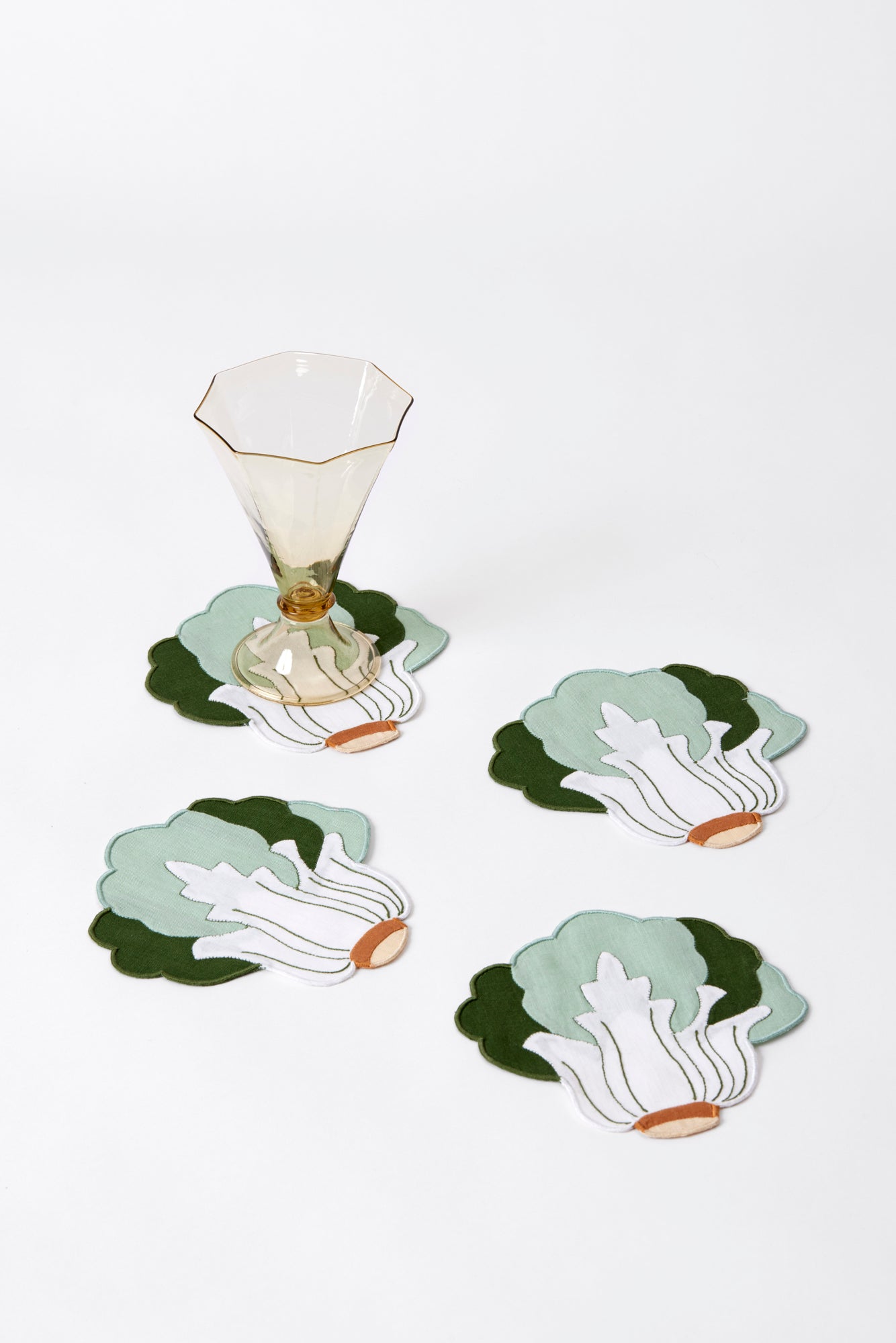 4 linen bok shoy inspired cocktail napkins on a white background. Linen cocktail napkin in the shape of a head of bok choy. Napkin is white center with green leaves and a brown base, along with embroidered details. On top of one of the napkins is a smoky colored hand blown glass goblet.