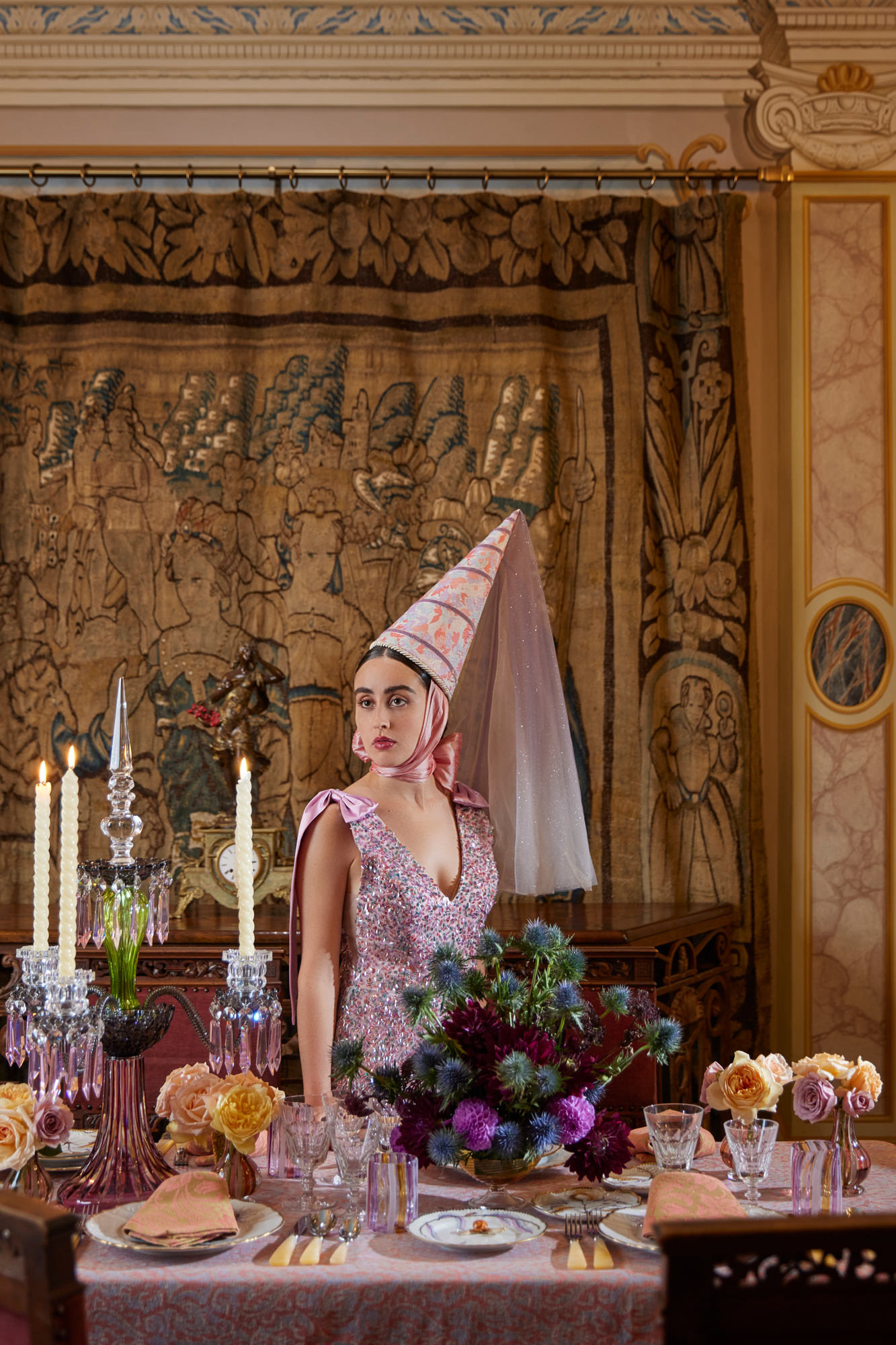 Scene of a girl standing at a dinner table. Girl is dressed in a sparkly gown with a hennin hat. In front of her is a table setting with an elegant glass candelabra with ivory candle tapers, a bouquet of thistles and fresh flowers, fine china settings, mother of pearl silverware, and hand blown glass tumblers.