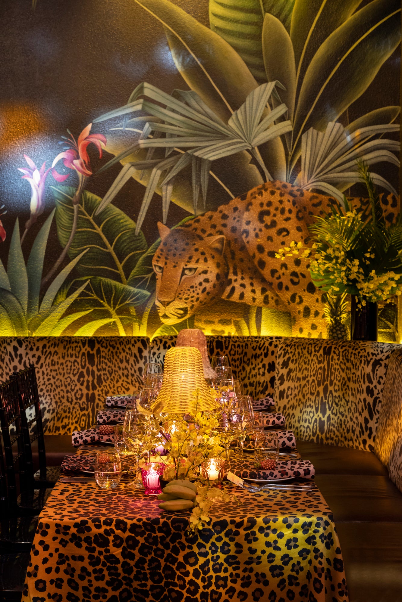 Table setting in a tropical lounge. Setting includes floor length leopard tablecloth, fine dishes and glassware, and wicker table lamps. Table surrounded by tropical florals and artwork. Large mural of a leopard on the wall behind the table.