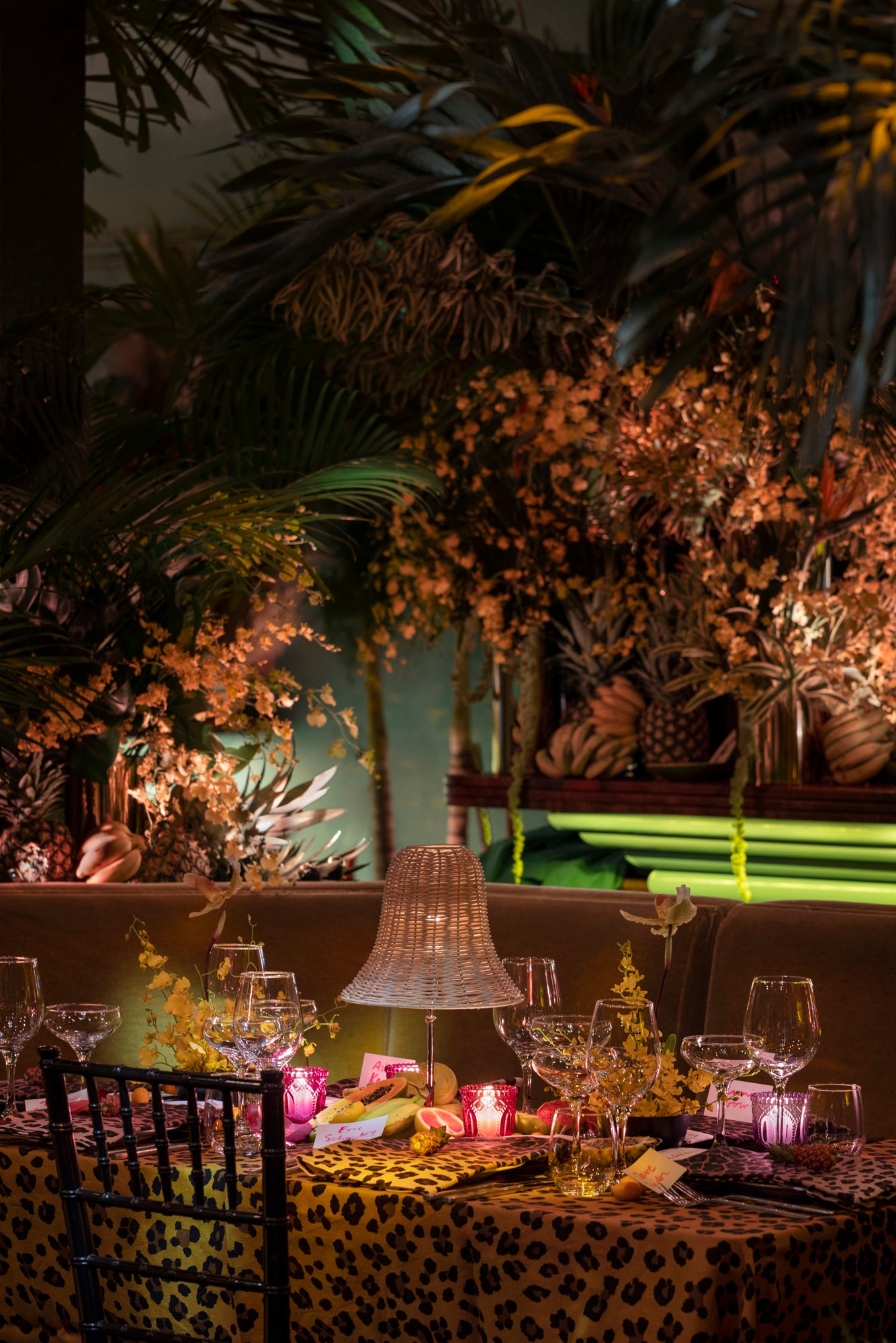 Table setting in a tropical lounge. Setting includes leopard tablecloth, fine dishes and glassware, and a wicker table lamp. Table surrounded by tropical florals.