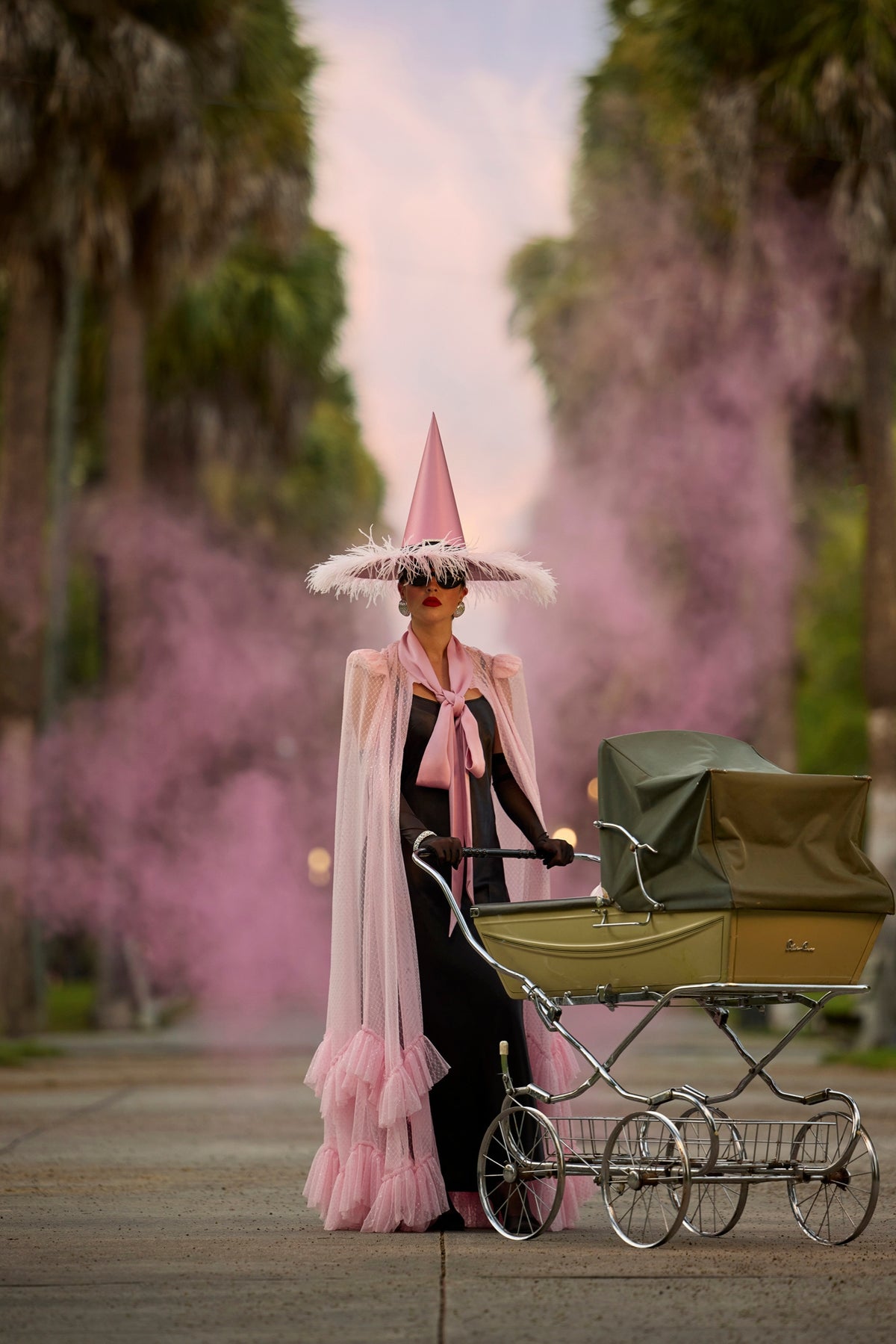 A person in a pink witch hat, pink cape, and black dress stands beside a vintage-style baby carriage on a path lined with palm trees and pink smoke.