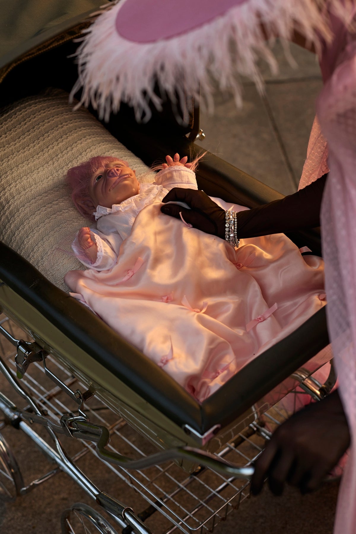 A baby werepup dressed in a light pink satin gown and matching pink hat lies in a black baby carriage, with an adult in pink clothing and white feather trim attending to the infant.