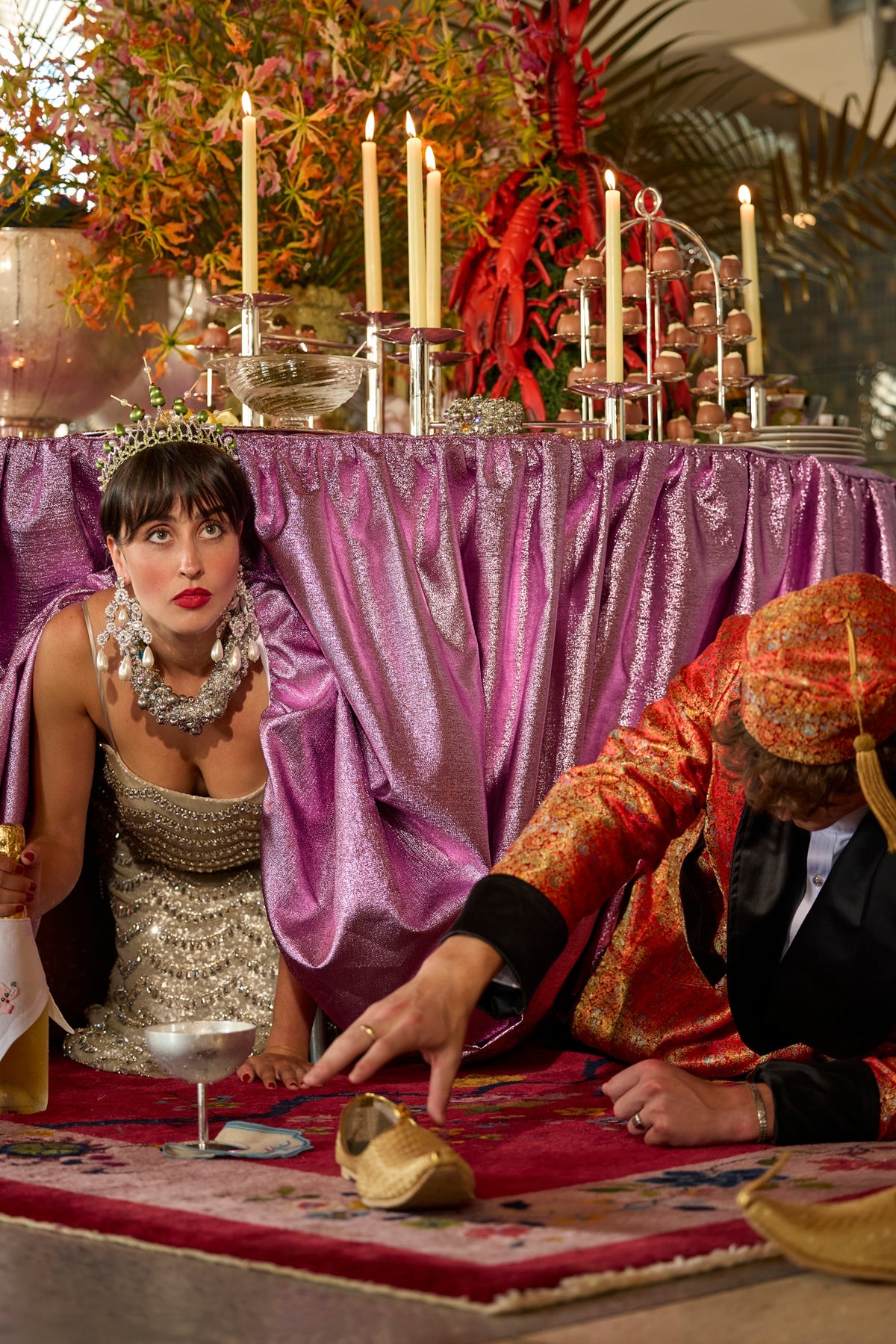 Woman in a silver crystal-and-sequin gown and jeweled tiara crouches beneath a purple-draped buffet beside a man in a red brocade jacket, with champagne and gold slippers nearby.