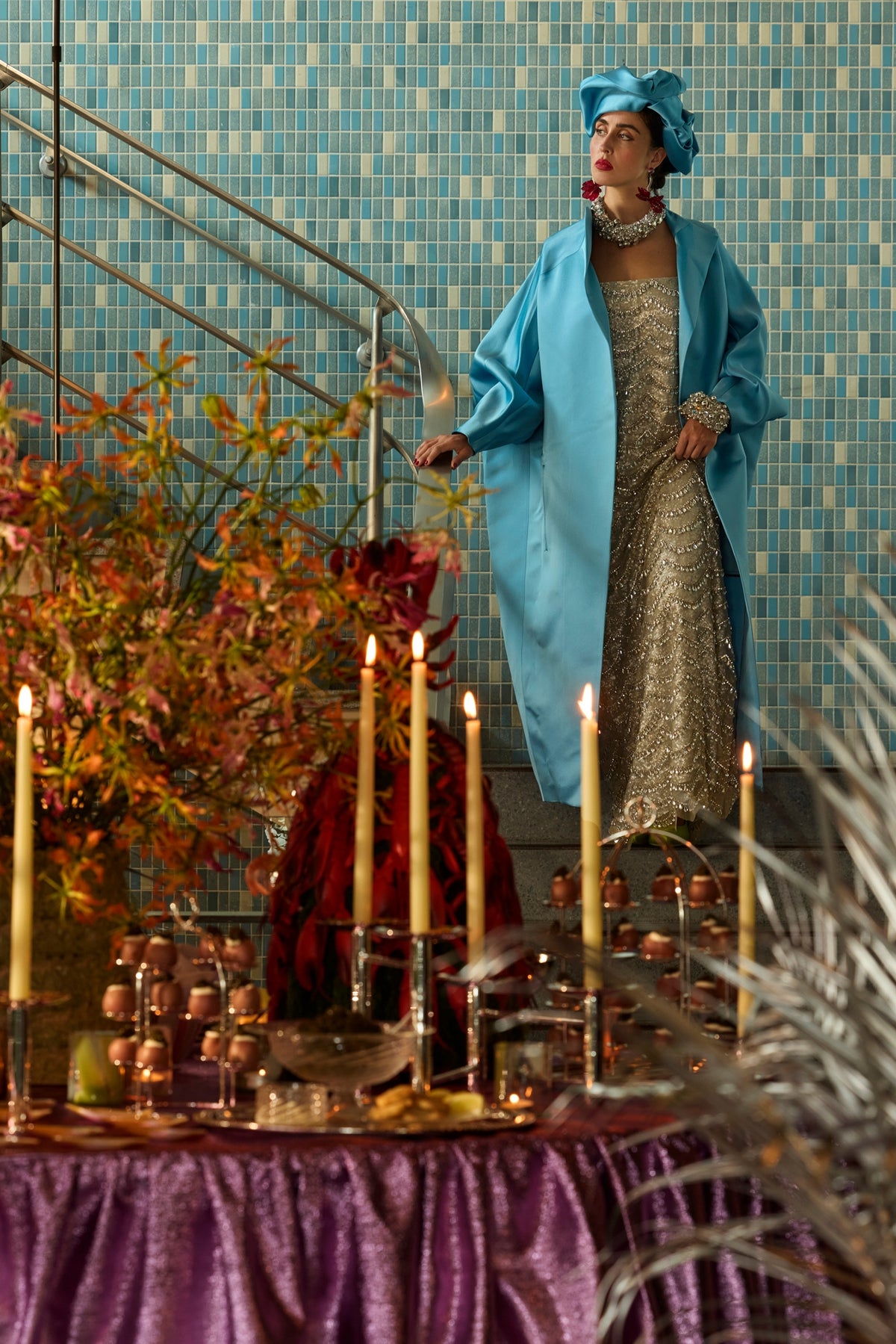 Woman in a silver crystal-and-sequin gown, turquoise satin coat and matching turban stands on a tiled staircase above an elaborate candlelit holiday buffet.