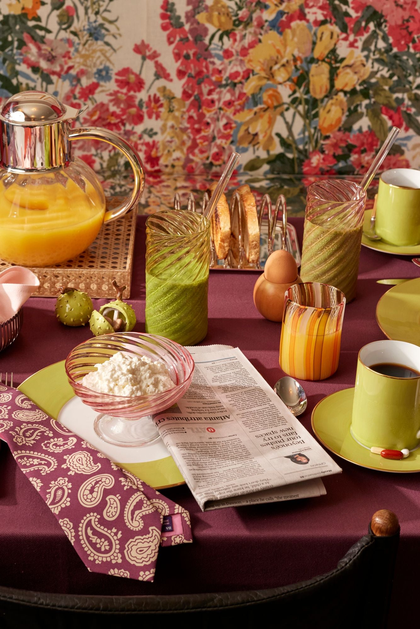 Table setting with orange juice, green smoothie, and coffee on a floral-patterned wall background.