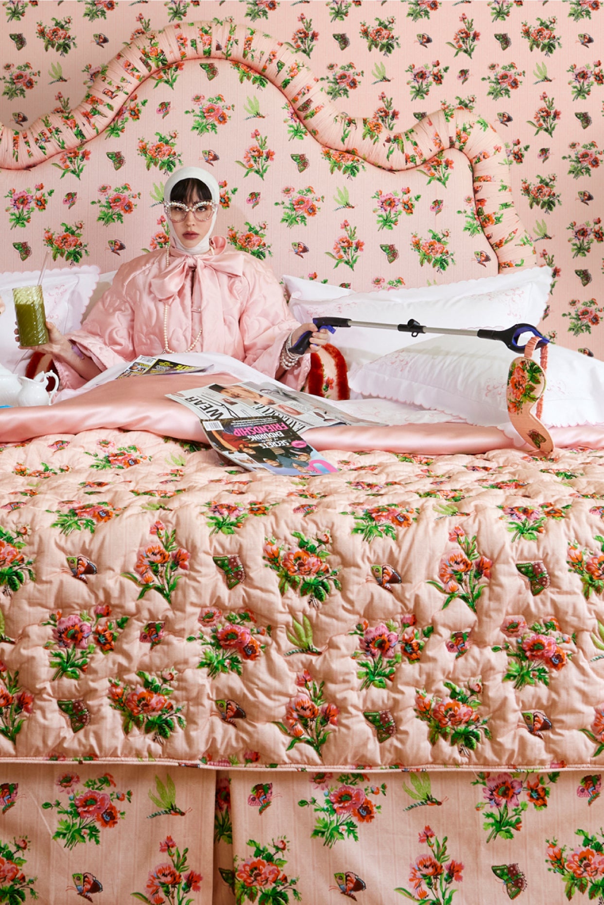 Woman sitting on a bed with floral bedding and headboard, holding a smoothie and a plastic reach tool.