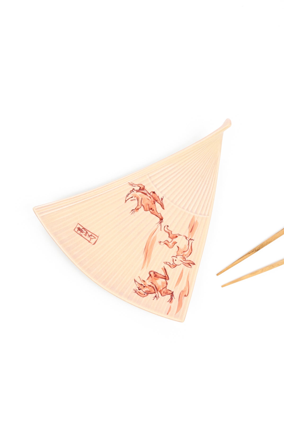 A ceramic plate in the shape of a traditional Japanese folding hand fan. Light blush in color with hand-painted frogs and rabbits, shown alongside wooden chopsticks on a white background.