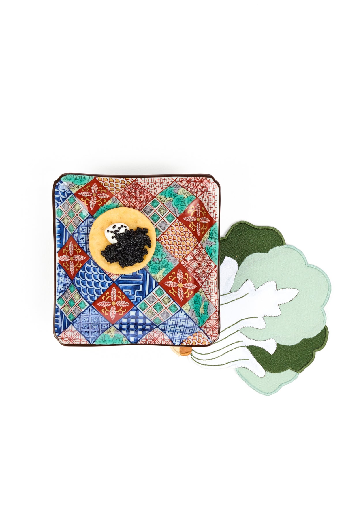 A square ceramic salad plate with a colorful patchwork tile pattern featuring red, blue, green, and orange geometric designs, set against a plain white background, topped with caviar and cremé frâiche. A light green and white bock choy shaped linen napkin is placed underneath to the lower right side. 