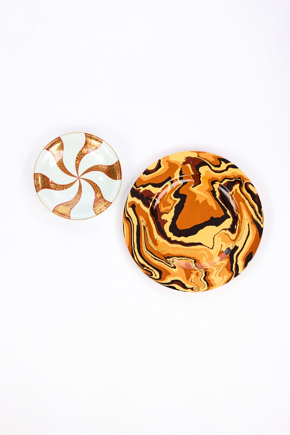 A round ceramic salad plate with a white background and a metallic gold and red spiral pattern, much like a peppermint, next to an orange, gold, and brown marbled ceramic plate.
