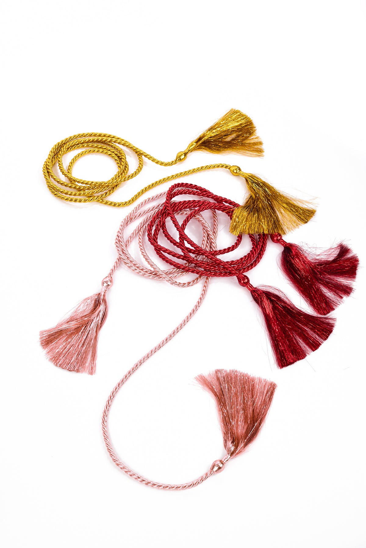 Three decorative braided tassels in gold, deep red, and light pink, arranged on a clean white background.