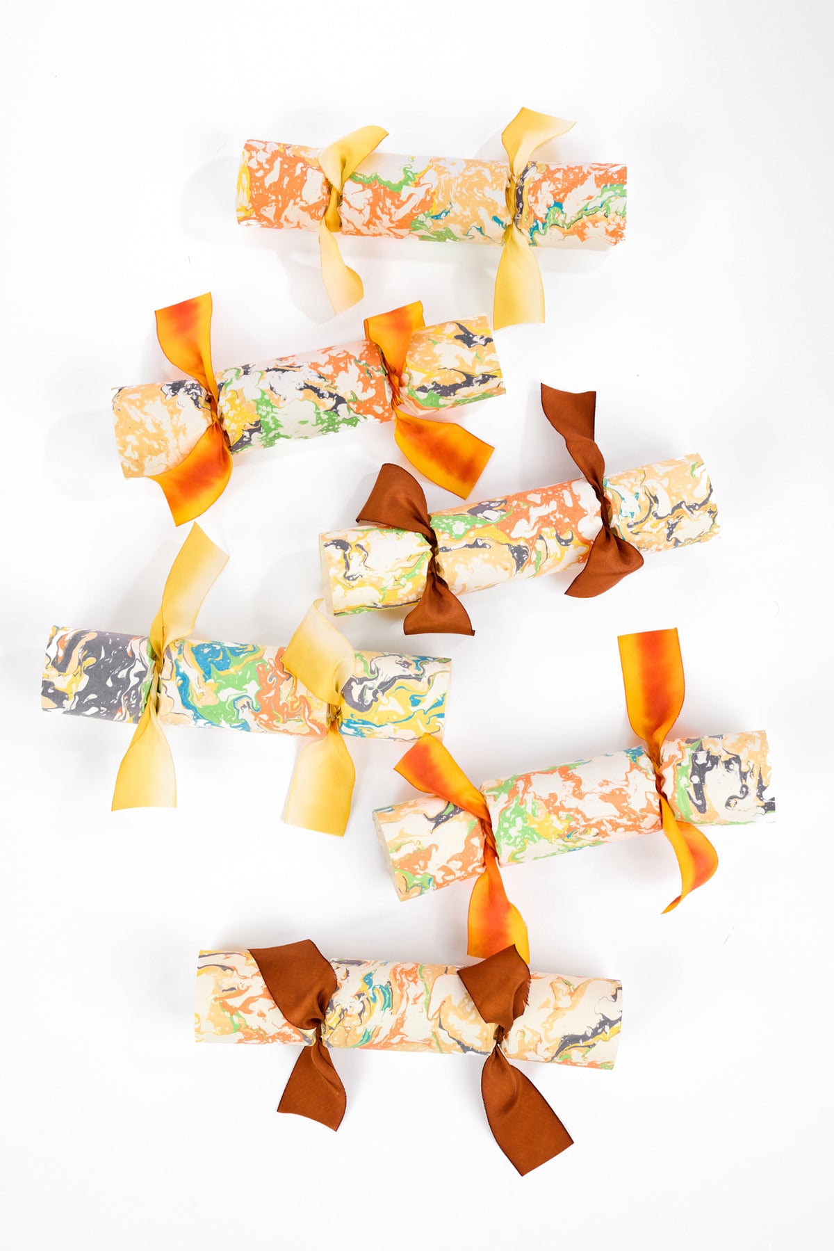 Nine Christmas cracker packs with marbled paper wrapping in autumn colors, tied with yellow and orange ribbons, arranged on a white background.