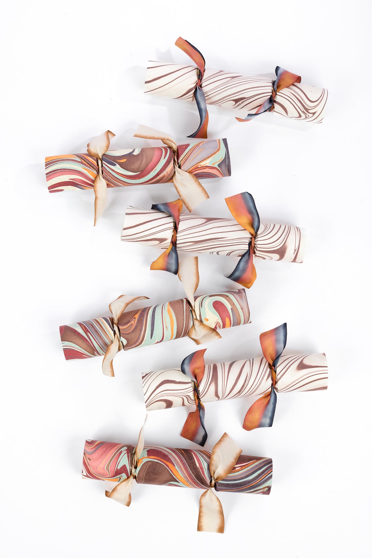 Five marbled paper Christmas crackers with brown, beige, orange, and blue swirls, tied with thin twine and ribbons, arranged vertically on a white background.