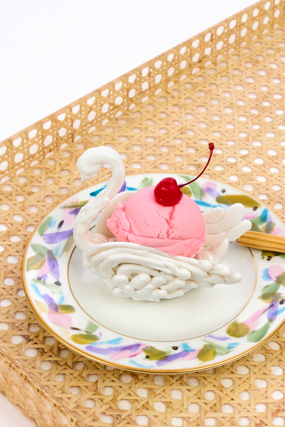 Faux porcelain swan made to look as if it was made of meringue holding a scoop of pink ice cream with a cherry on top atop a patterned abstract plate with a gold rim. All sitting on a beige caned tray.