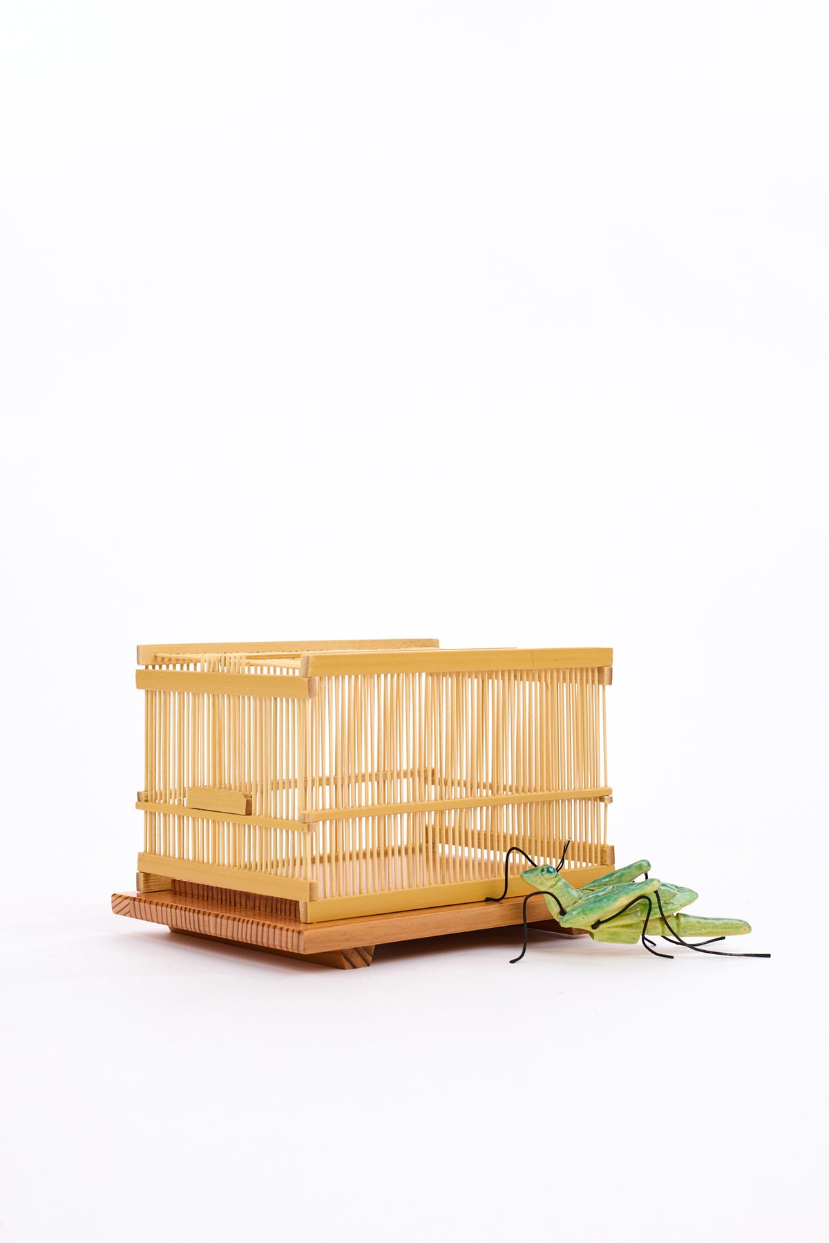 Traditional bamboo Japanese cage atop a wooden slab with a green ceramic grasshopper against a white backdrop.