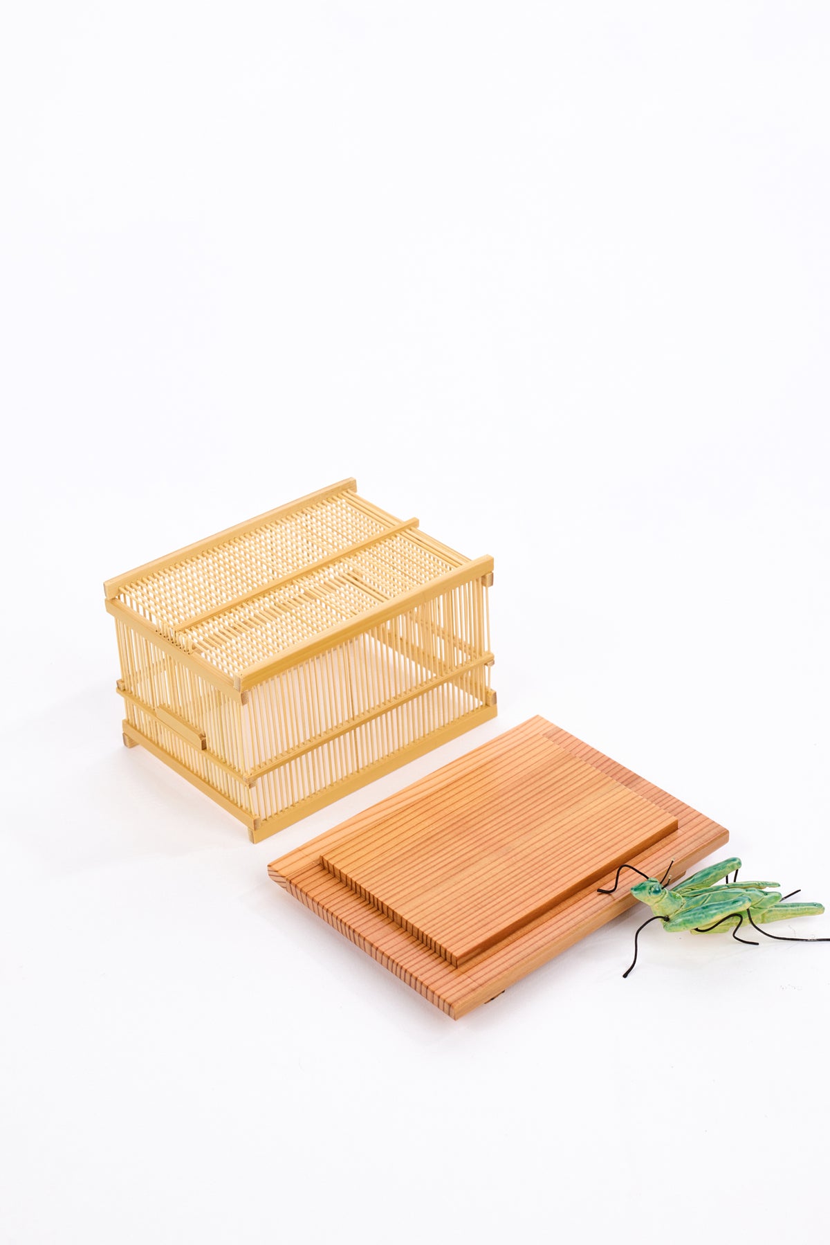 A miniature wooden cage with a woven lid, alongside a matching wooden tray or cover and a small green ceramic grasshopper decoration, photographed on a clean white background.