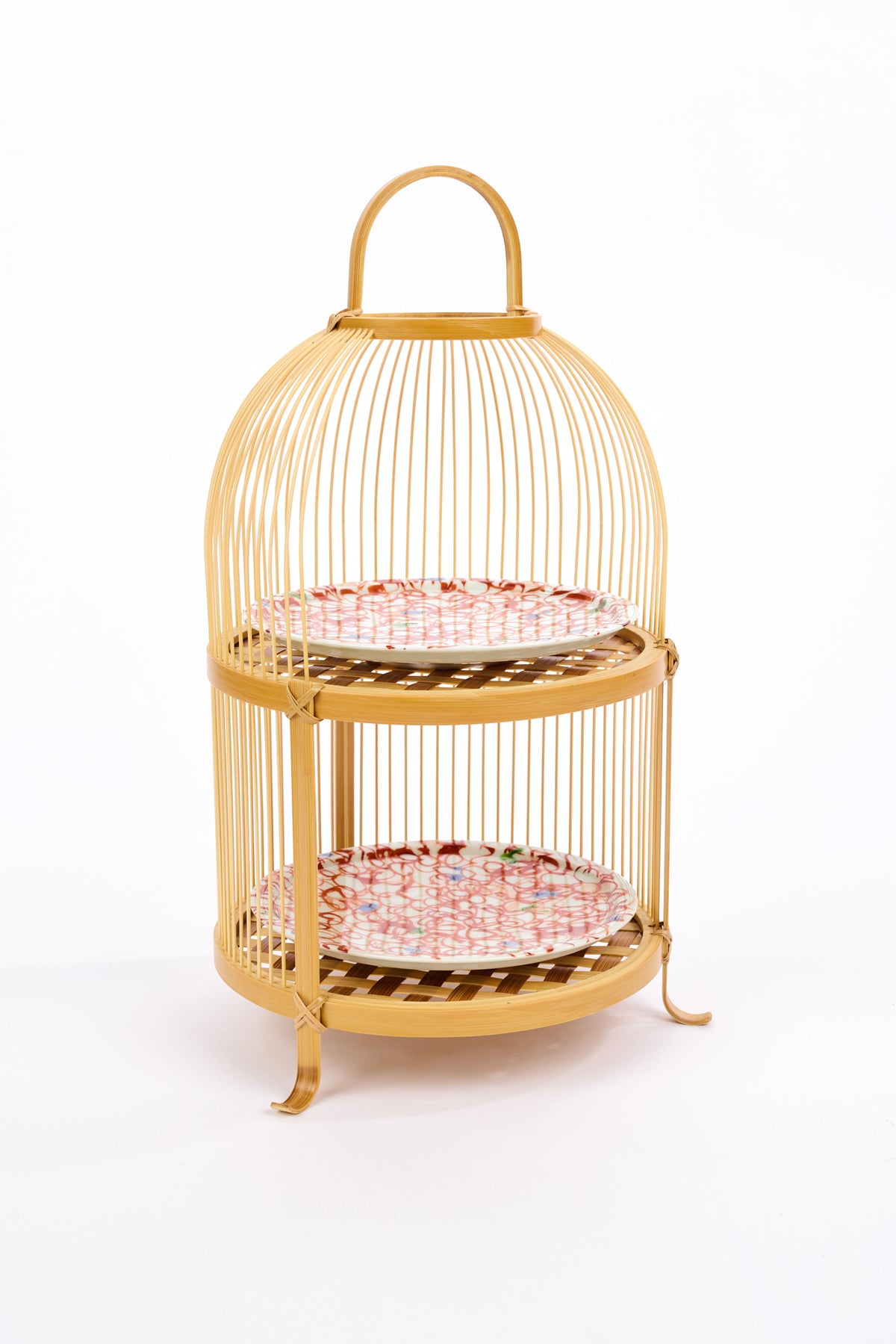 Two-tiered bamboo tea stand with woven shelves holding abstract red patterned ceramic plates with blue and green dots against a white backdrop.