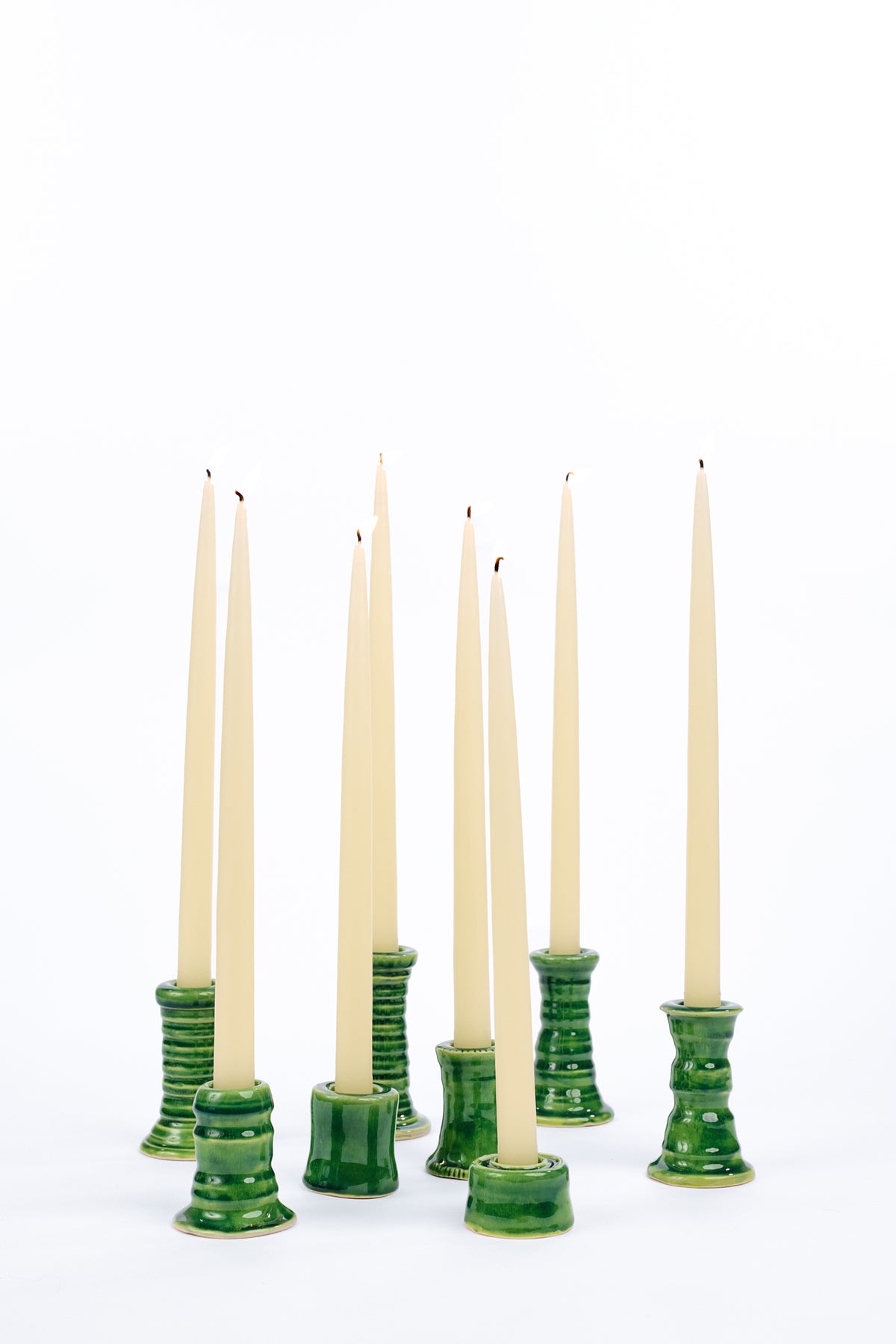 A set of eight taper candles in cream-colored glass holders with glossy green bases, arranged on a seamless white background.