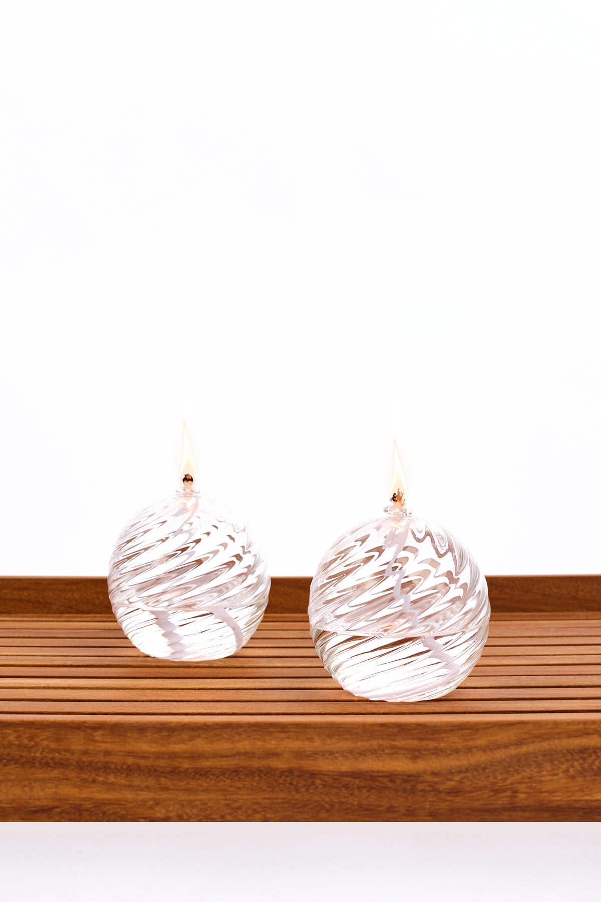 Two decorative spiral glass oil lanterns with a burning wick, both clear, displayed side by side on a wooden slatted surface against a white background.