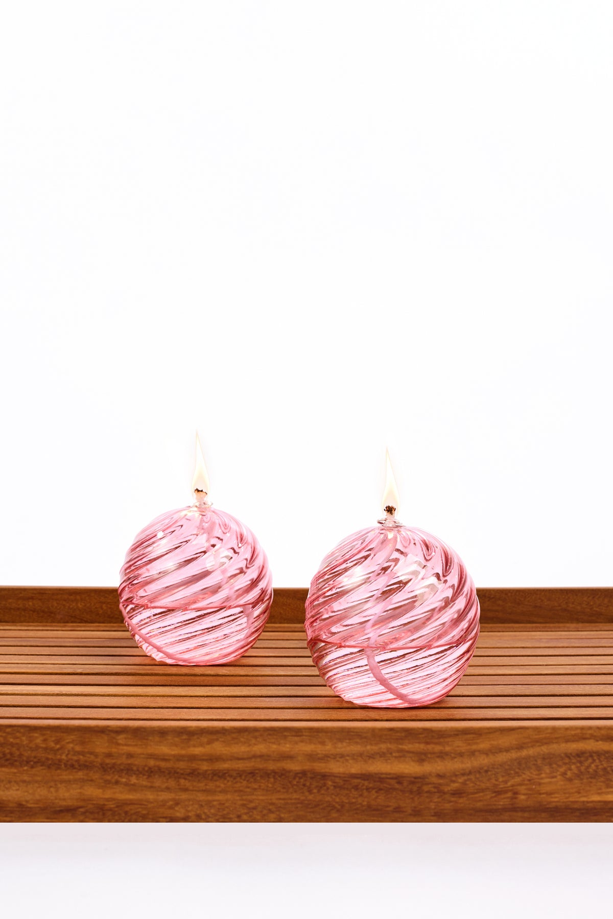 Two decorative spiral glass oil lanterns with a burning wick, both pink, displayed side by side on a wooden slatted surface against a white background.