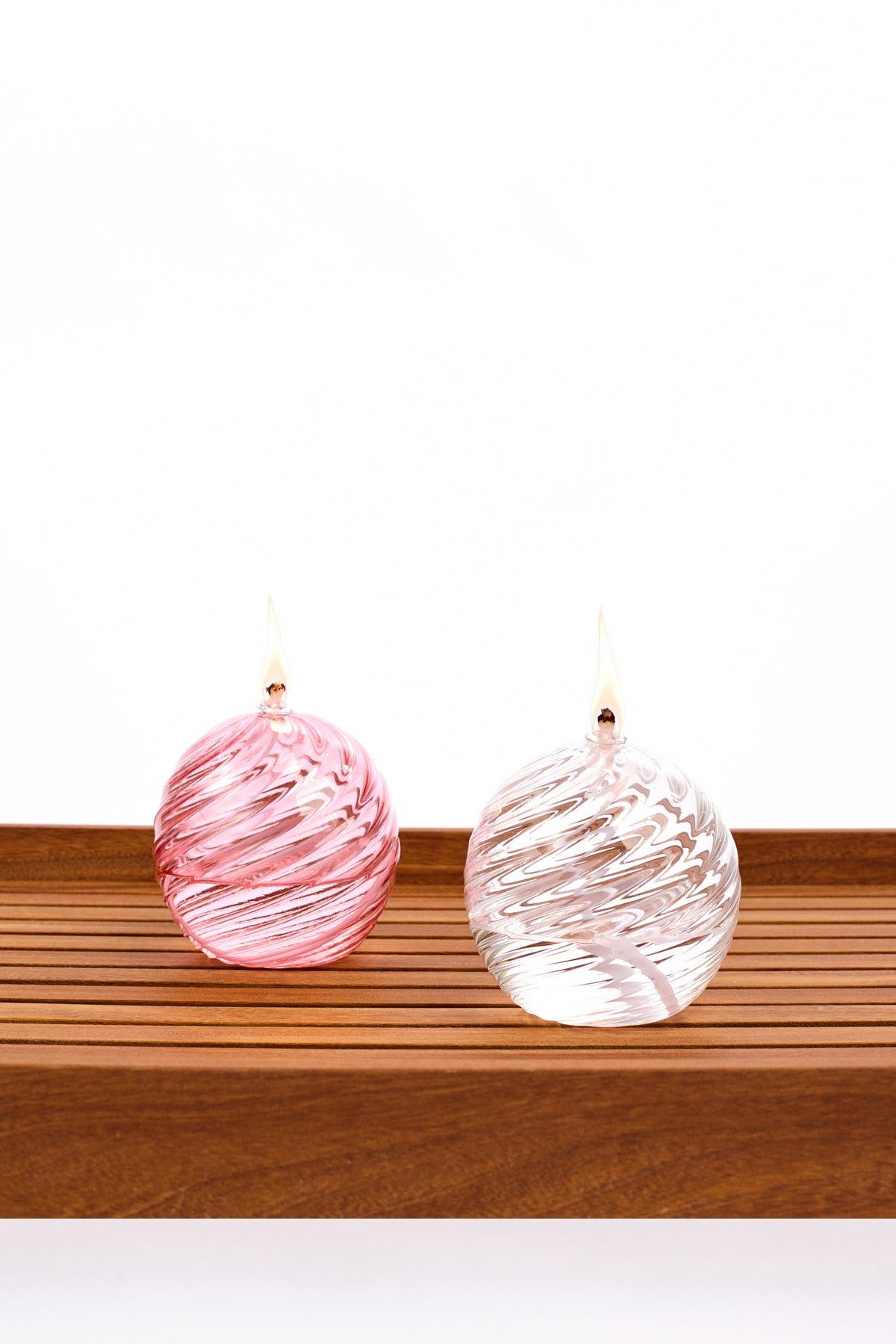 Two decorative spiral glass oil lanterns with a burning wick, one pink and one clear, displayed side by side on a wooden slatted surface against a white background.