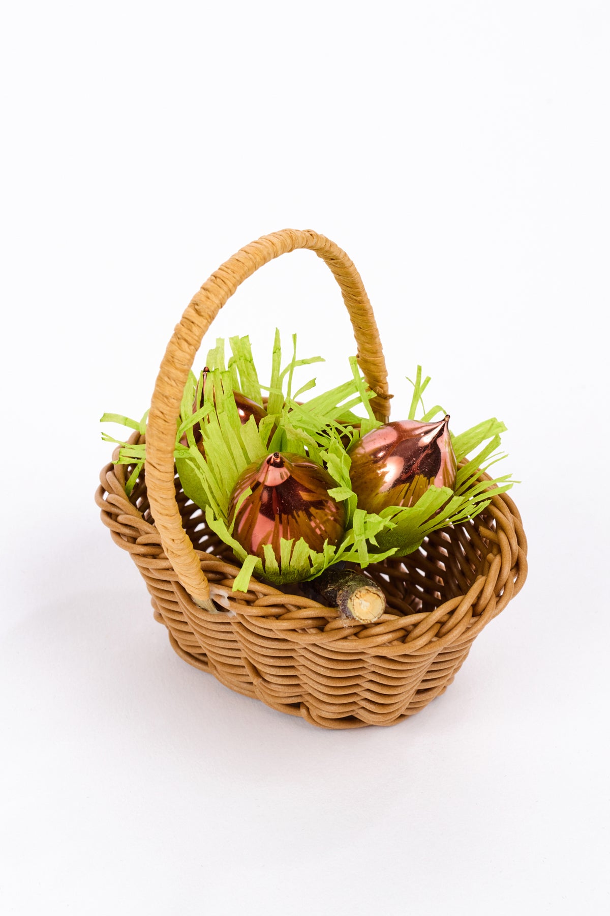 A small woven wicker basket filled with bright green artificial grass and shiny metallic hazelnuts, photographed on a clean white background.