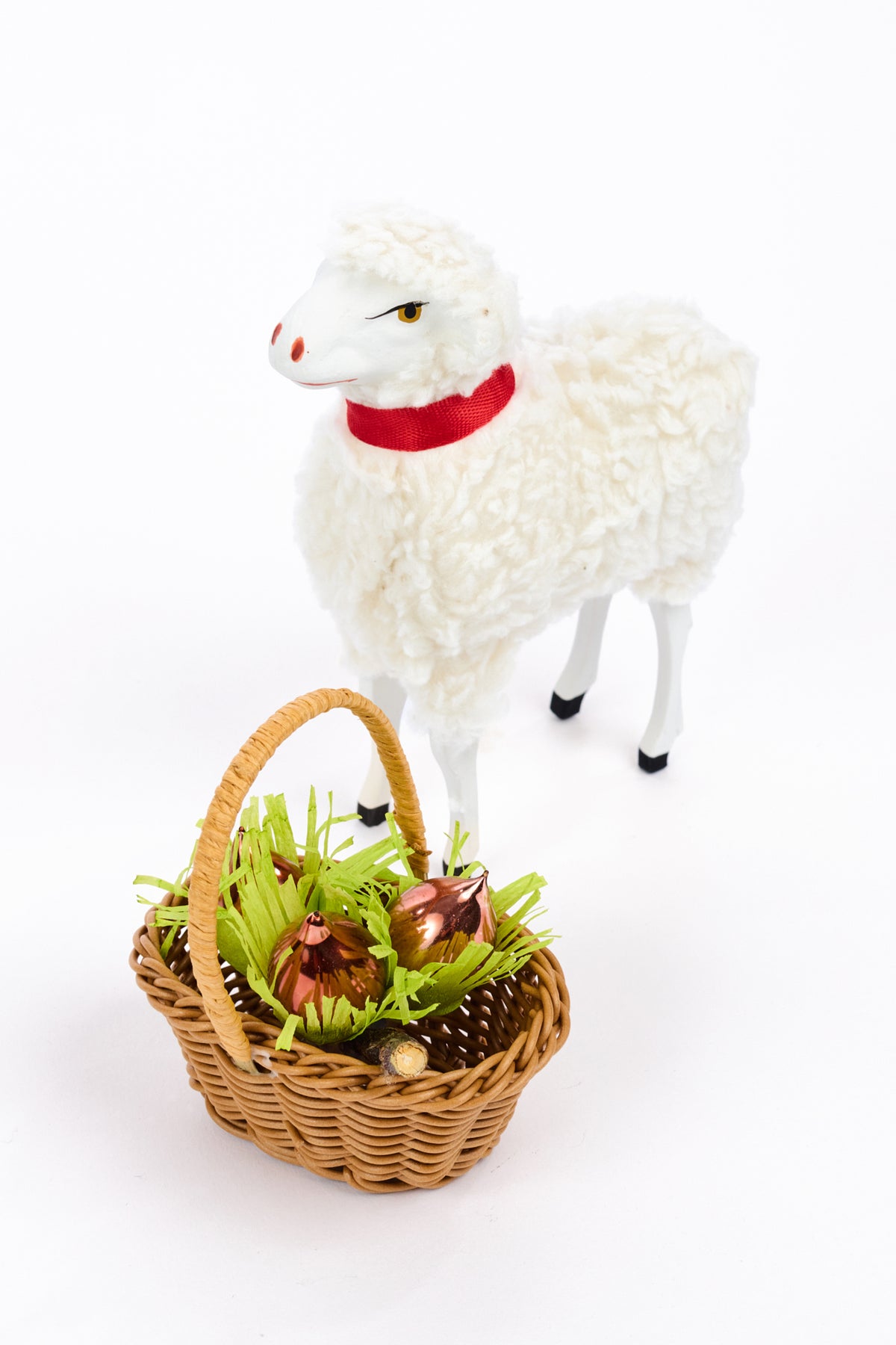 A white plush sheep figurine wearing a red collar stands beside a small woven basket filled with green grass and decorative brown hazelnuts on a clean white background.