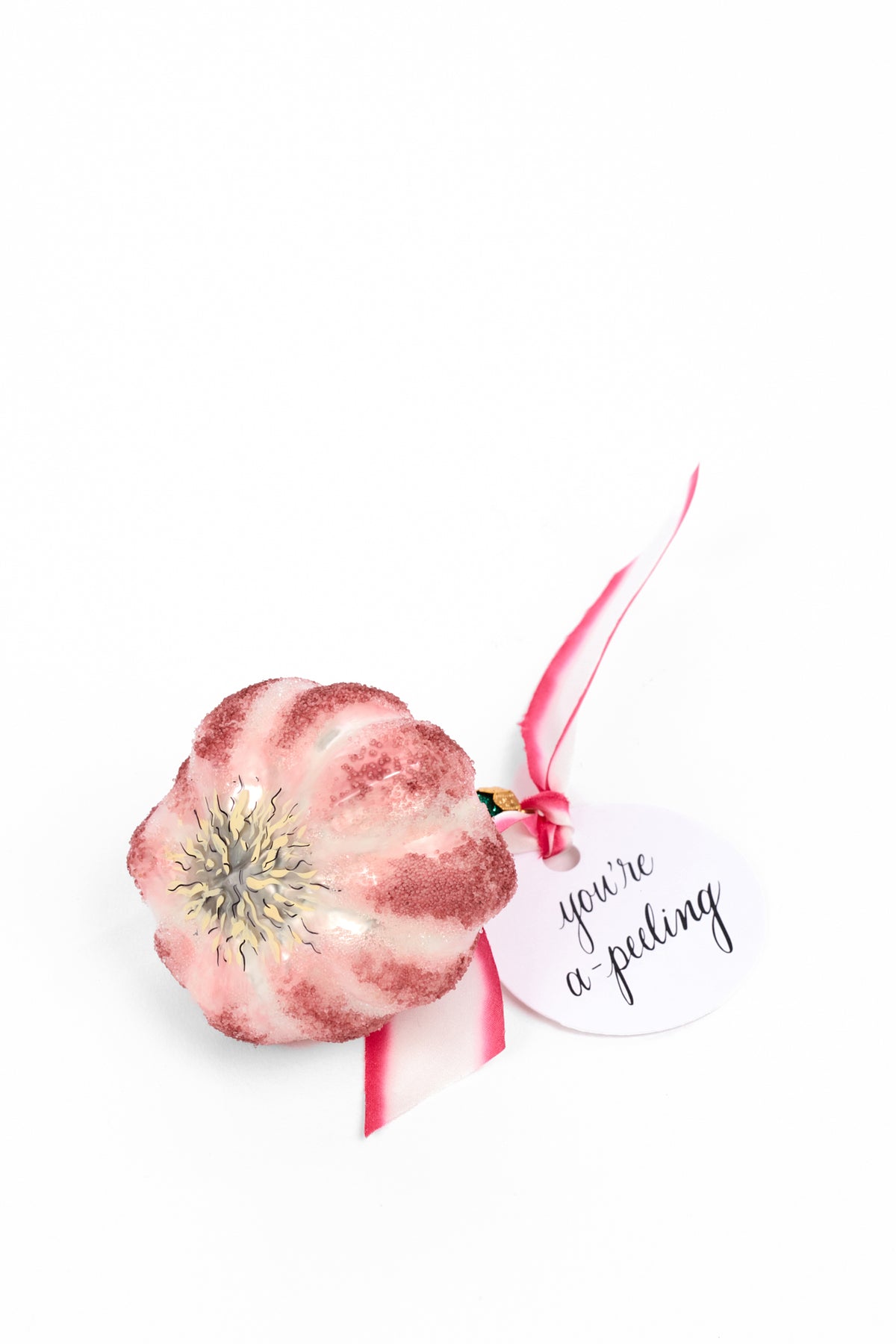 A small decorative ornament shaped garlic covered in pink and white glitter, with a pink ribbon and a round white tag that reads 'you're a-peeling'.