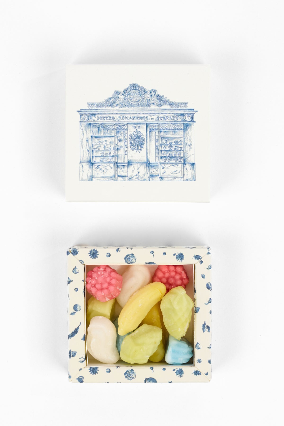 A top-down view of a square gift box set on a white background. The lid is white with a blue architectural illustration, and the open box below contains several pastel-colored, raspberry-shaped candies or soap bars in pink, yellow, green, and blue, set in a white box with a blue floral border.