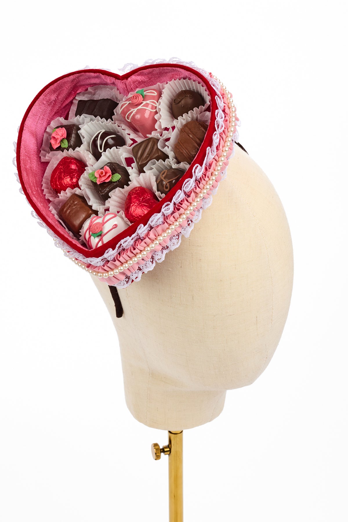 A fascinator in the style of a heart-shaped box of assorted chocolates displayed on a cream-colored mannequin head against a white background.