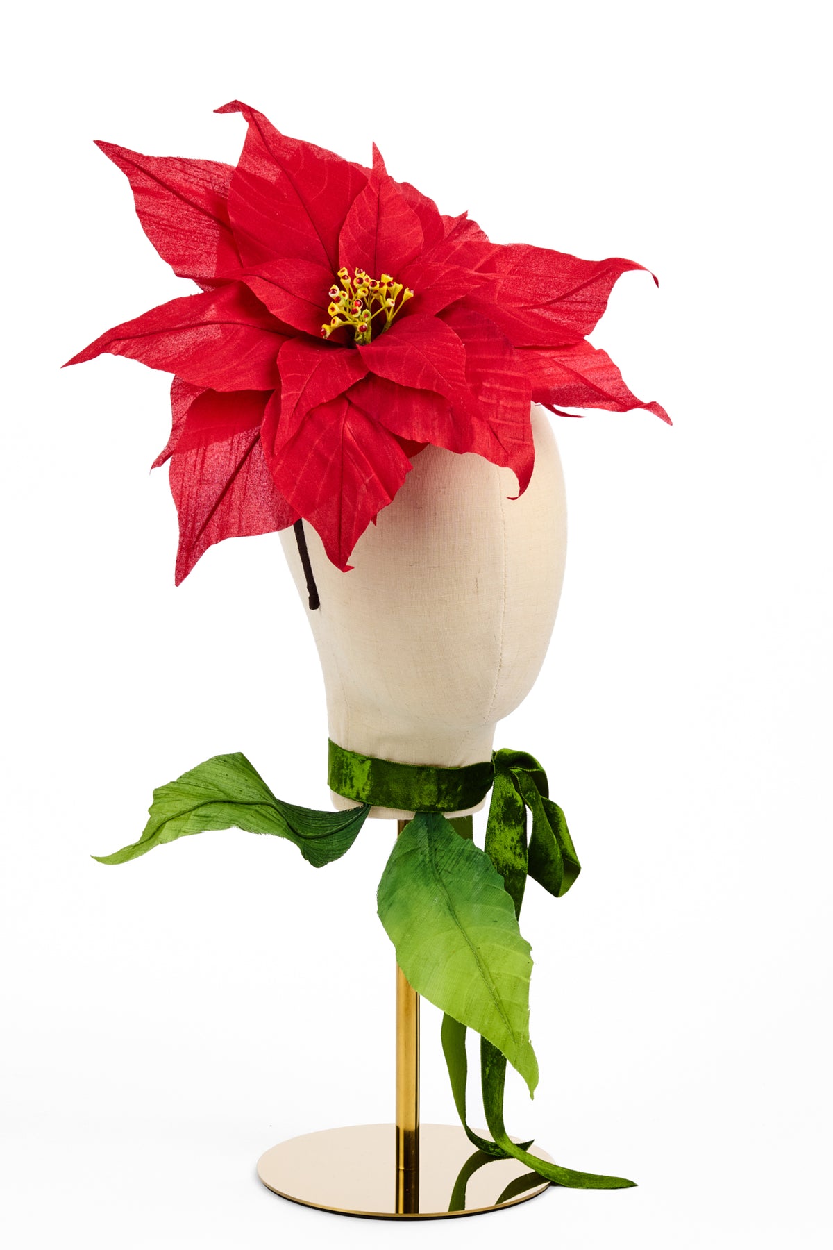 A red poinsettia artificial flower with green leaves and a yellow center, displayed on a cream mannequin head with a green ribbon and gold stand against a white background.