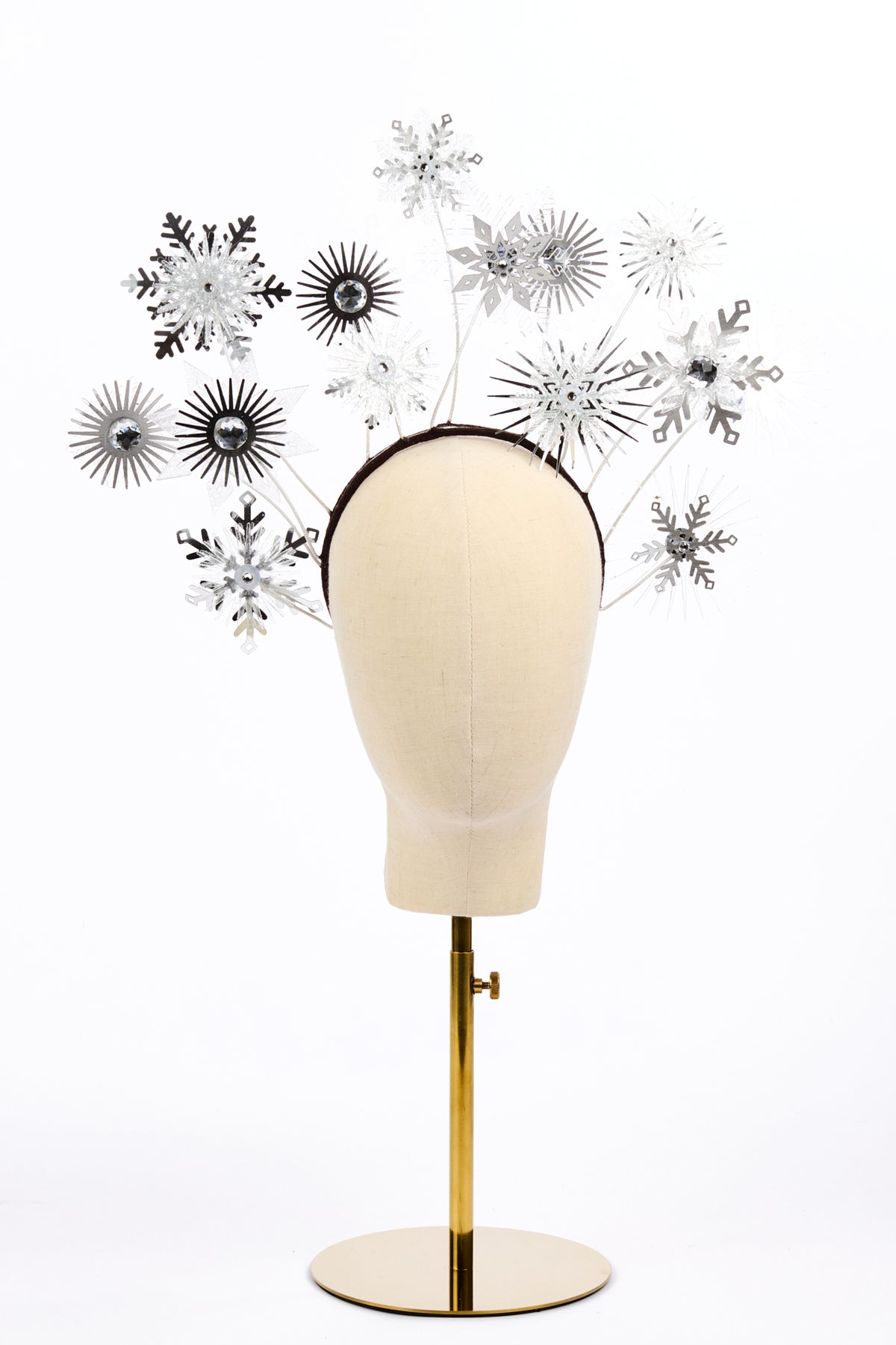 A silver and white snowflake and sunburst headpiece displayed on a cream fabric mannequin head mounted on a gold stand, against a plain white background.
