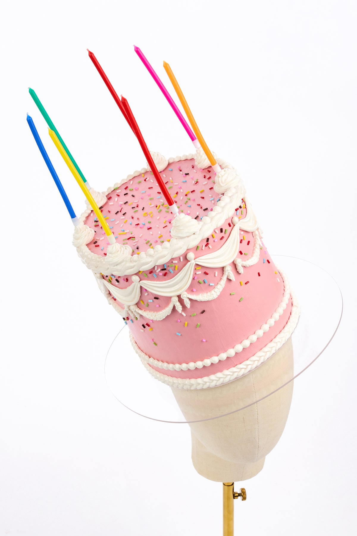 A pink birthday cake with white frosting piped borders and colorful birthday candles inserted into the top, displayed on a mannequin head against a white background.