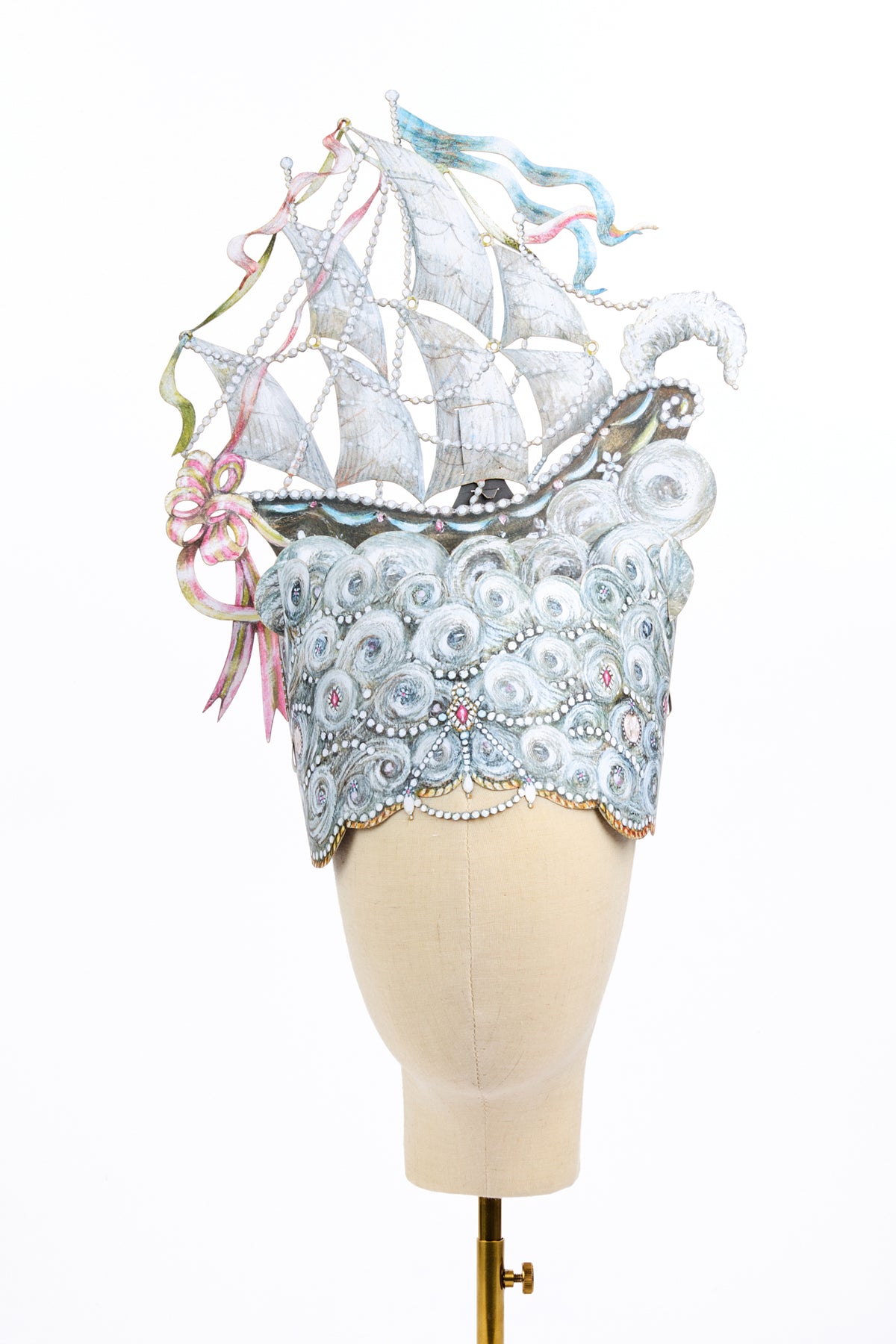 Sculptural headpiece depicting a black-and-white sailing ship with pearl-trimmed sails, blue and pink ribbons, and a white plume. The ship rises from silver waves decorated with pearls and colorful jewels on a cream mannequin head.