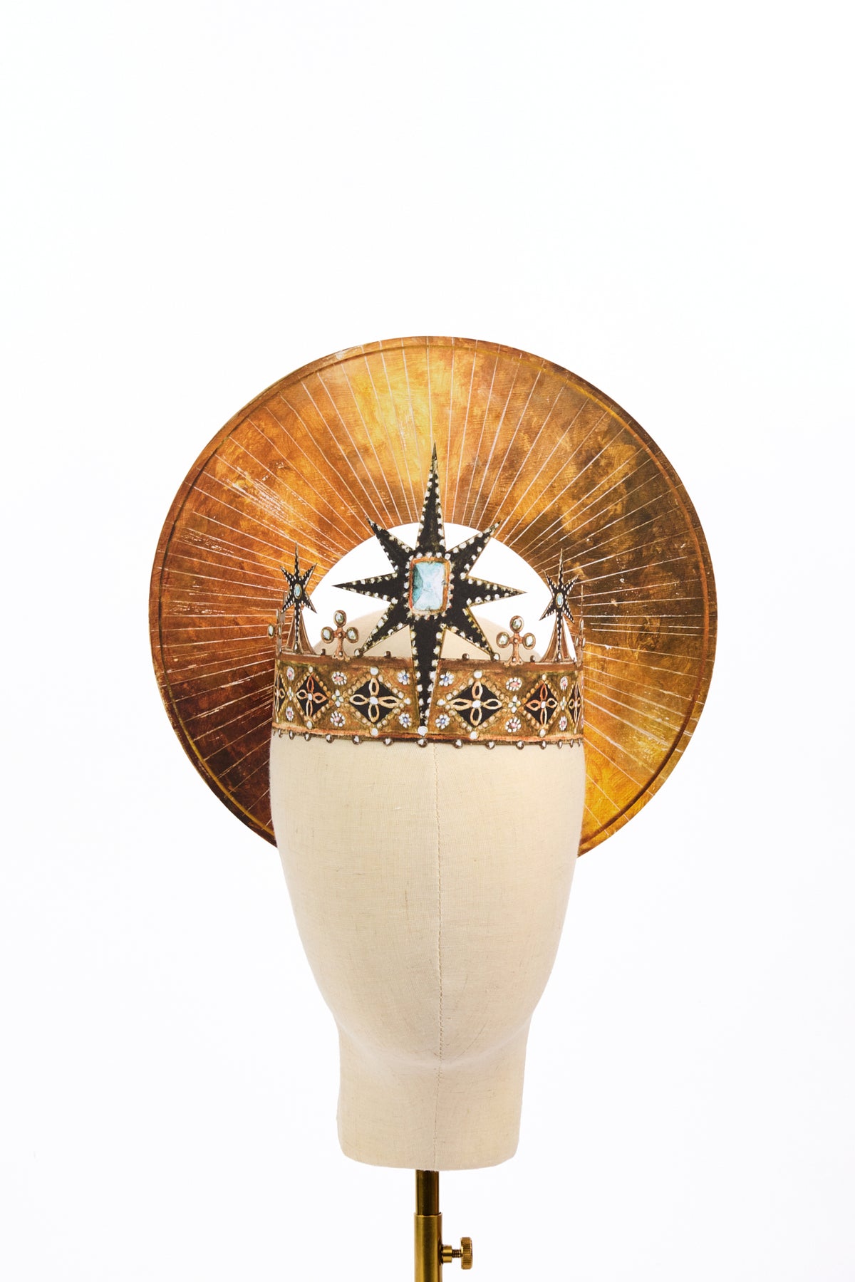 Ornate gold crown decorated with black starbursts, pale blue stones, pearls, and floral motifs. A large circular gold halo with radiating lines rises behind the crown on a cream mannequin head.