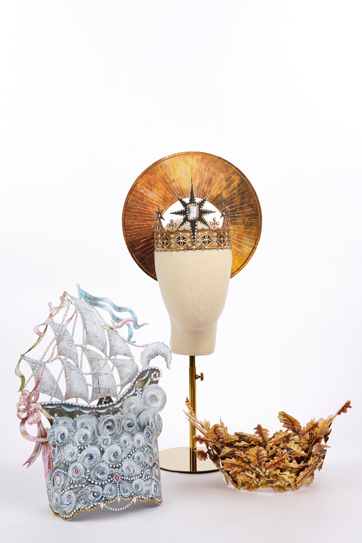 Three elaborate headpieces displayed together on a white background: a sailing ship rising from jeweled silver waves, a gold crown backed by a circular sunburst halo, and a separate gold crown formed from overlapping oak leaves and acorns.
