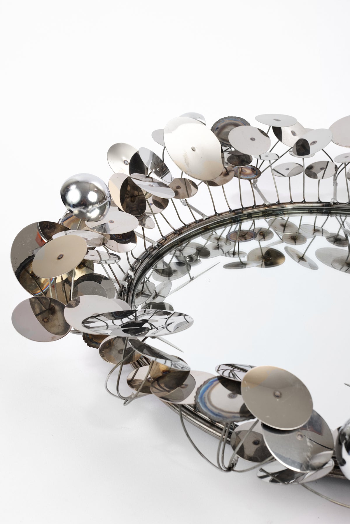 A modern sculptural wall mirror with a reflective silver frame made of numerous overlapping circular metal discs and thin wire supports, photographed on a seamless white background.