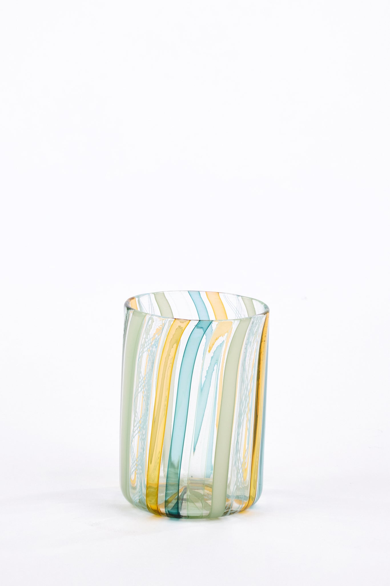 A clear glass tumbler with vertical hand-blown stripes in light blue, yellow, and pale green, set against a seamless white background.