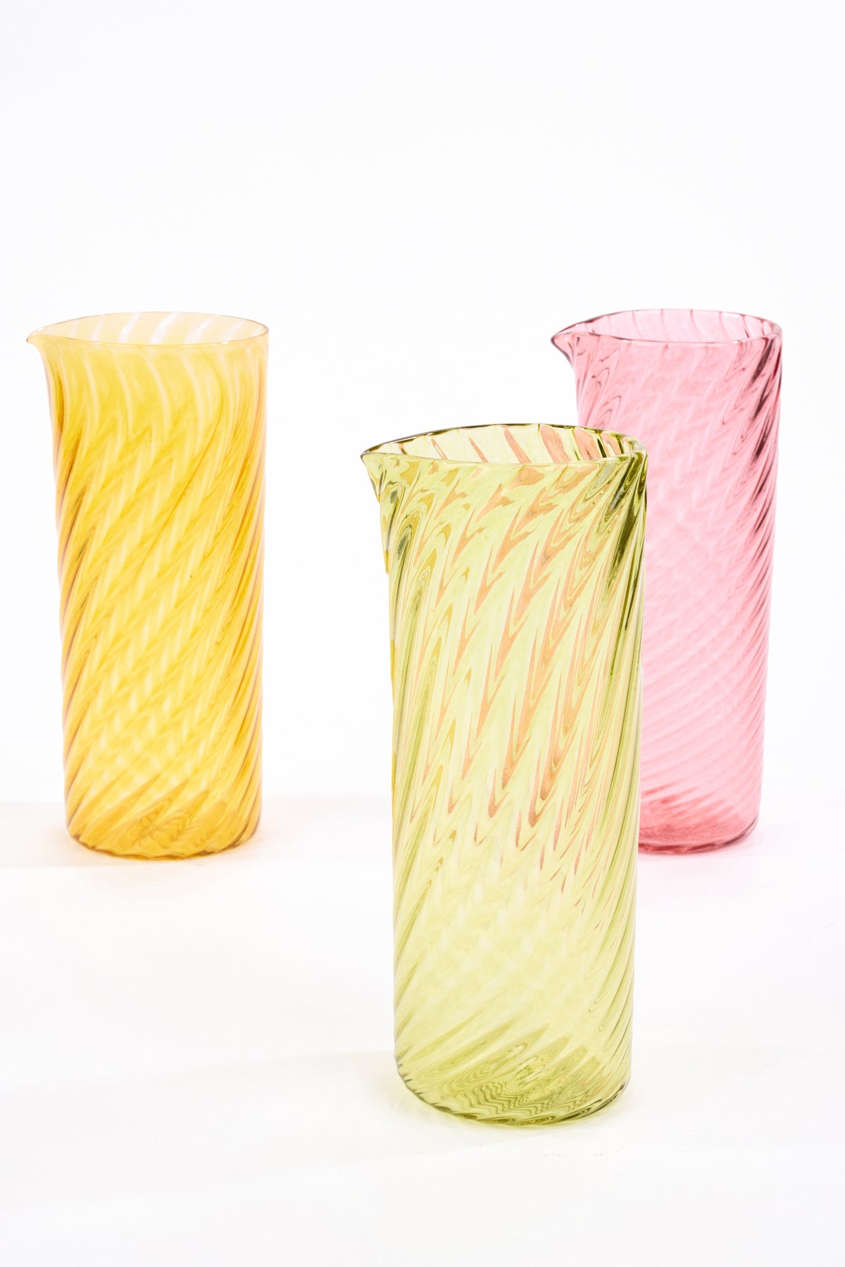Three tall, twisted glass pitchers in yellow, light green, and pink, arranged on a seamless white background.