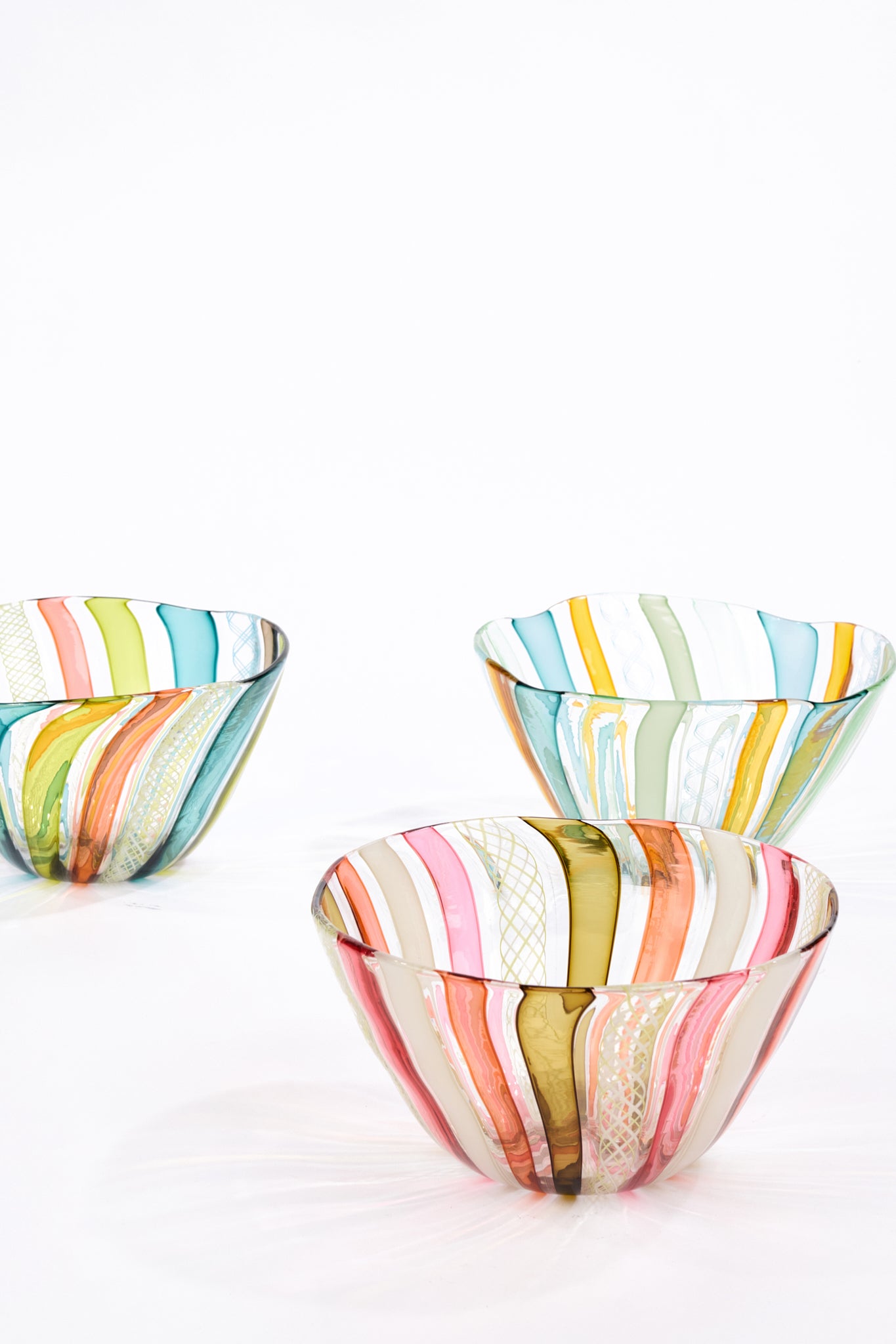 Three hand-blown glass bowls with colorful vertical stripes in shades of pink, orange, yellow, green, blue, and white, arranged on a clean white surface against a white background.