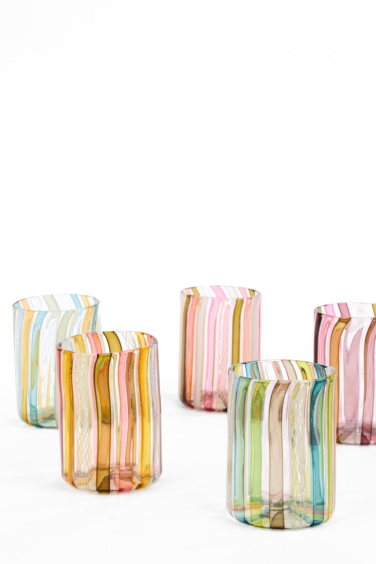 Five small cylindrical glass tumblers with vertical multicolor stripes in pastel and warm tones, arranged on a clean white background.