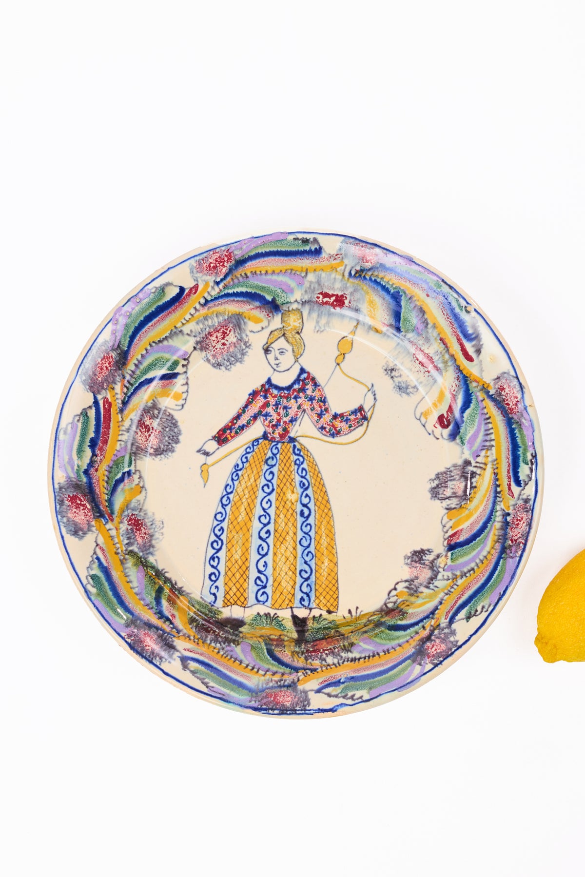 A round ceramic plate with a hand-painted traditional Italian style illustration of a woman in a colorful dress, surrounded by a vibrant border of blue, yellow, green, and pink floral motifs, set against a plain white background with a small yellow lemon visible on the right edge.