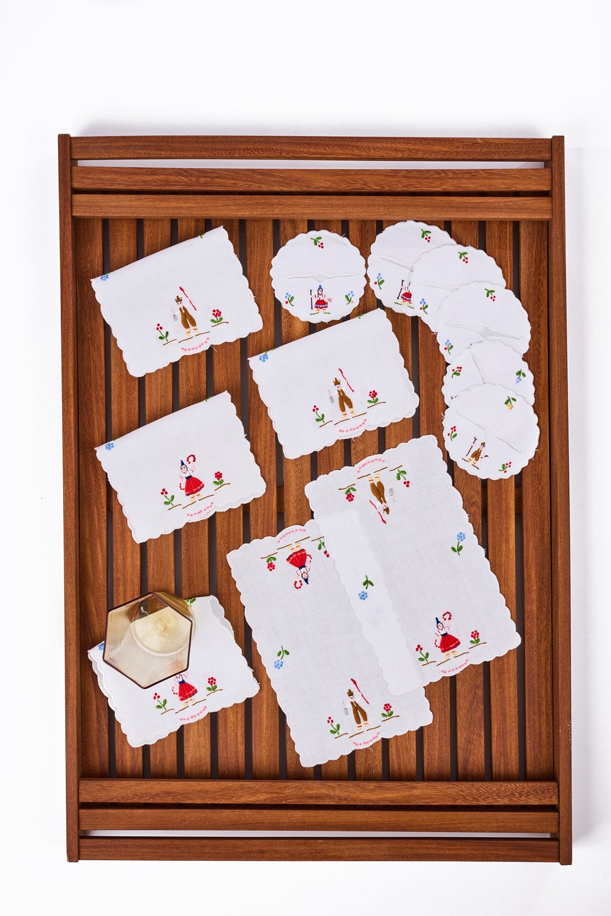 A wooden slatted serving tray holding a coordinated set of white embroidered napkins, round coasters, with a small hexagonal glasss on a white background.