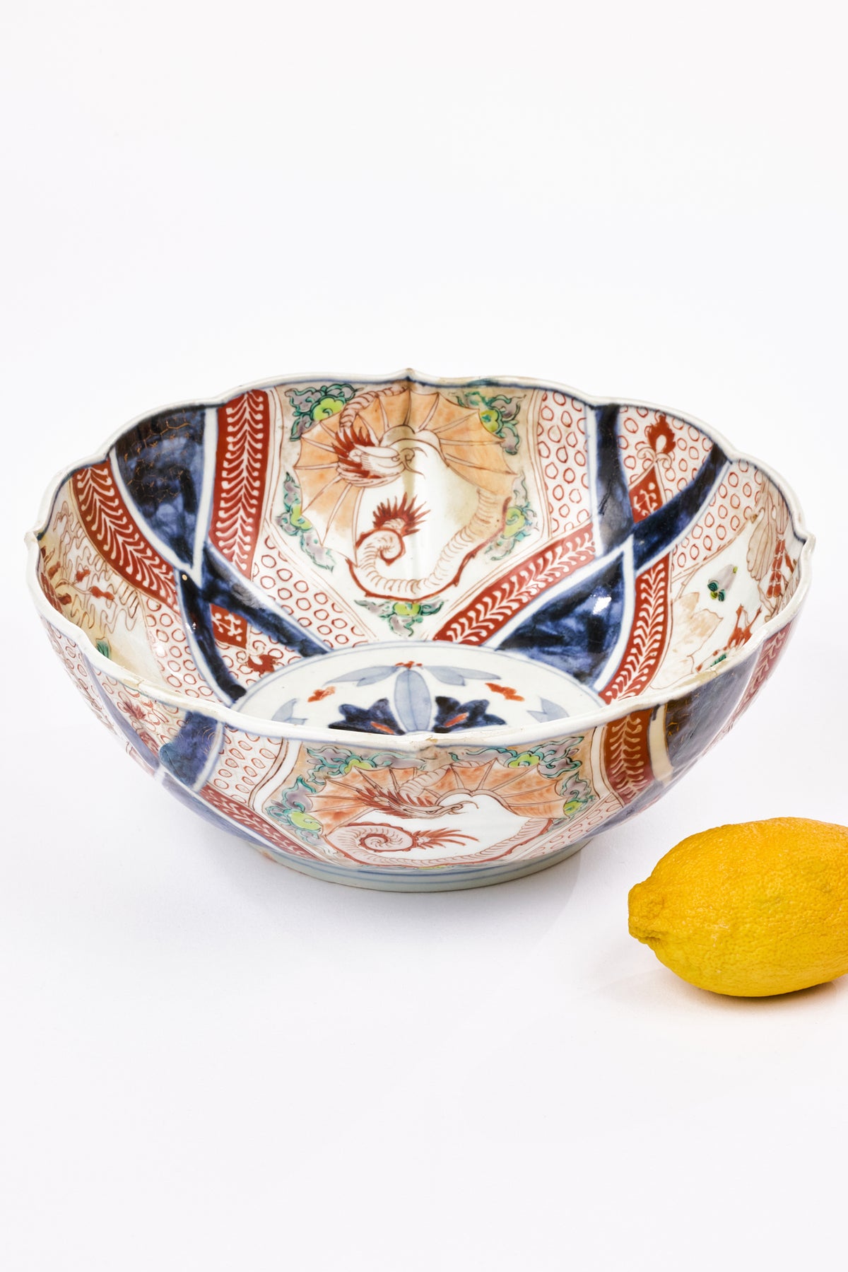 An antique Japanese Imari ceramic bowl with scalloped edges, featuring hand-painted floral and geometric patterns in blue, red, green, and beige on white porcelain. A fresh yellow lemon rests to the right of the bowl on a clean white background.